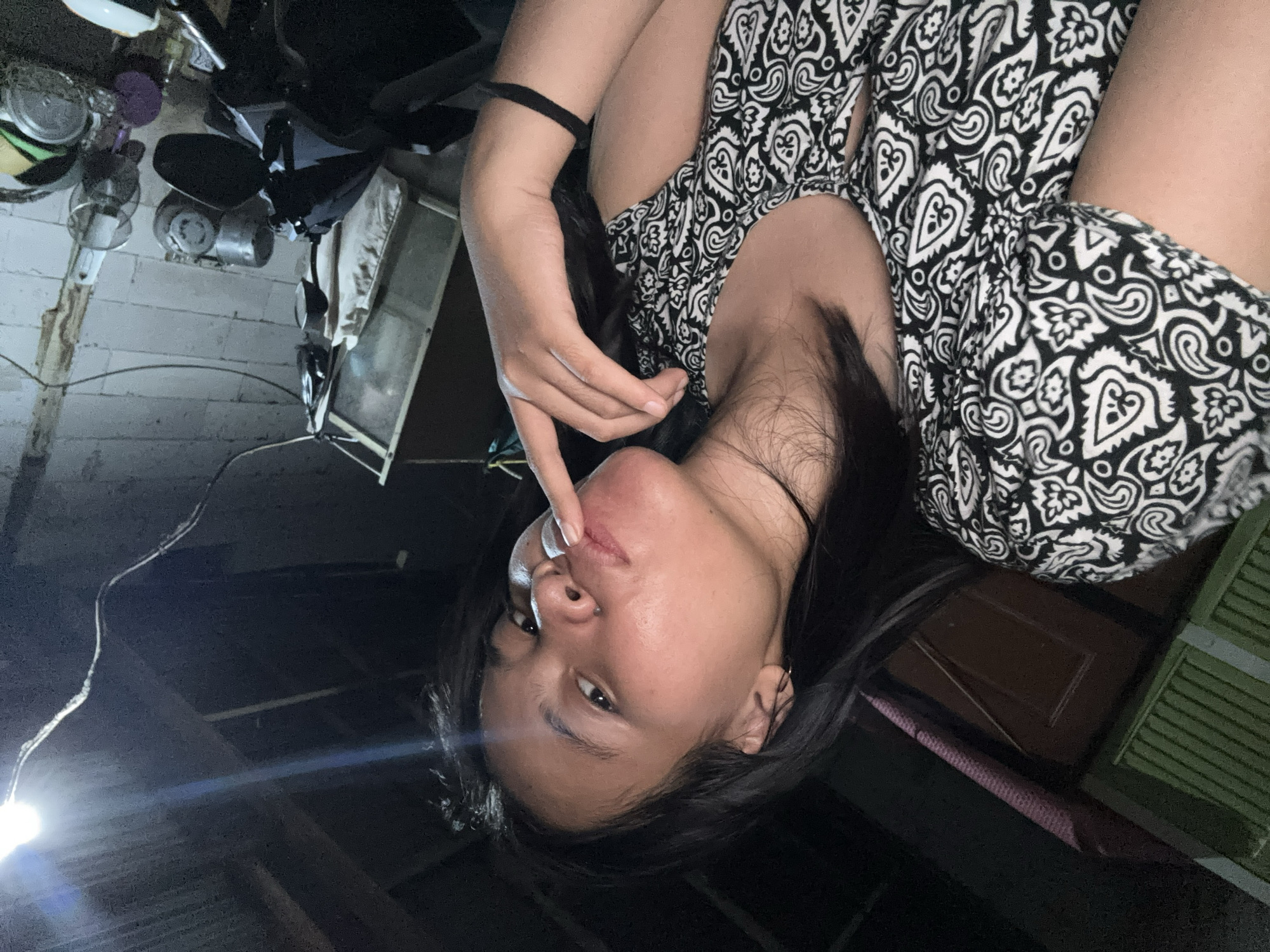 A young woman, with dark hair and brown eyes, poses for a selfie in what appears to be a cluttered, dimly lit interior space. She is wearing a black and white patterned dress. Her right index finger is touching her lips, and she is looking directly at the camera. Behind her, the environment is somewhat obscured by shadow but includes a wooden ceiling, exposed wires, a light bulb, and various objects on shelves or surfaces, suggesting a storage area or a workshop. The scene suggests an indoor residential or utility space in East Jakarta, Indonesia.