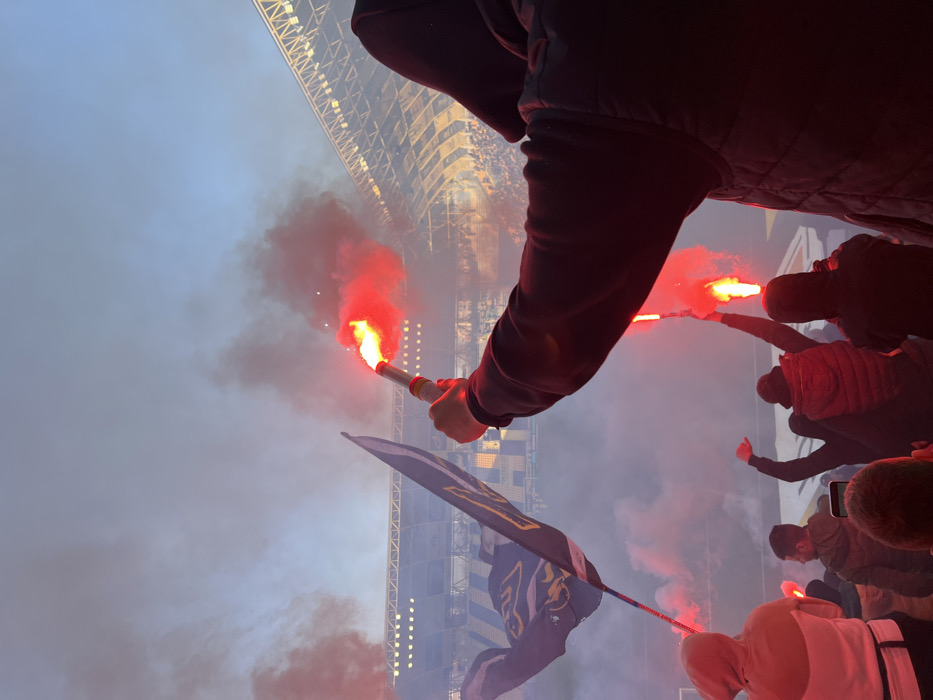 The image depicts a crowded football stadium at dusk, specifically Hristo Botev Stadium in Plovdiv, Bulgaria. In the foreground, a person viewed from behind wears a dark hooded garment and a dark quilted vest, raising their right arm to hold an active red flare. The flare emits intense red light and dense red smoke. To the immediate left of this individual, a dark-colored flag displaying a yellow stylized emblem, likely a lion, and the text "Botev Plovdiv" is held aloft on a pole.
In the midground, multiple other individuals are visible. At least two other people are observed from behind, each holding an active red flare with their arm raised. One person on the far left wears a light-colored hooded top. In the central foreground, the back of a head is visible, with a smartphone horizontally positioned at eye level, suggesting recording.
The background reveals the stadium's tiered seating, portions of which are yellow and dark-colored, filled with spectators. Overhead stadium floodlights are illuminated. The sky above is a fading twilight blue. Red smoke from the pyrotechnics permeates the entire scene, partially obscuring the stadium structure and the sky.