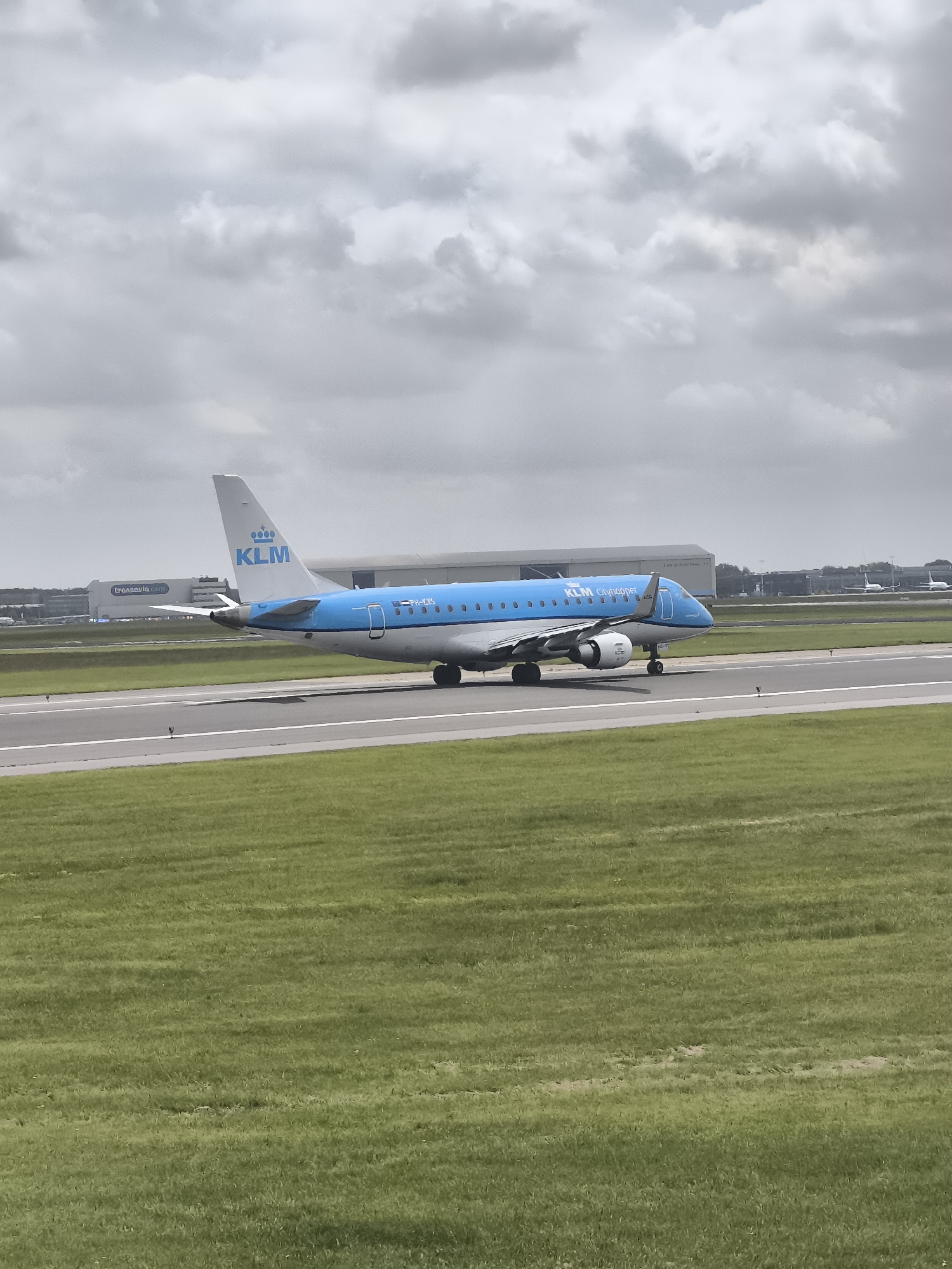 A KLM Cityhopper aircraft is on a runway. In the background are airport buildings. The sky is overcast. The location is Unknown, Unknown. The aircraft is light blue and white. The area adjacent to the runway is green grass.