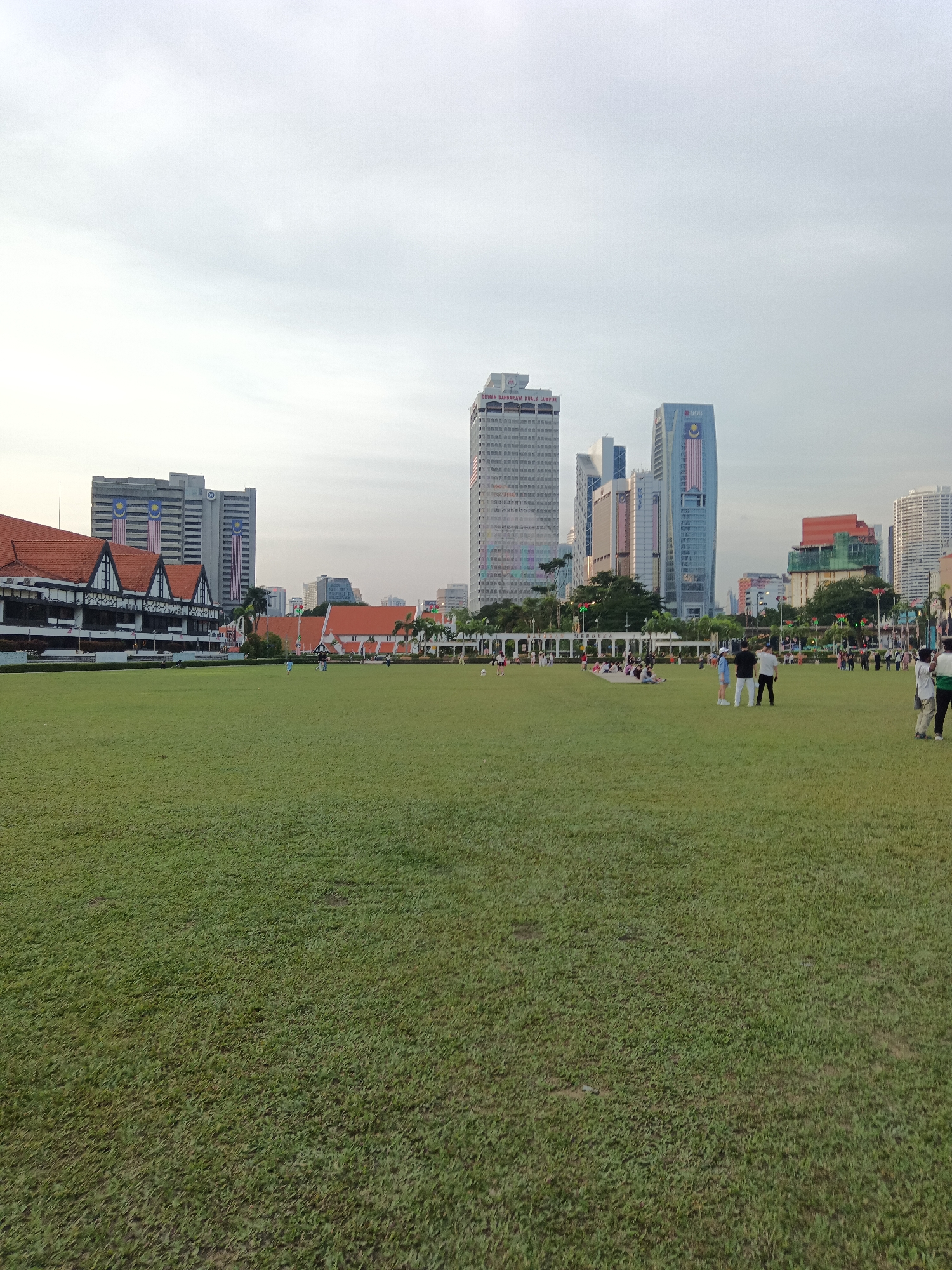 The image captures a wide-angle view of the Dataran Merdeka (Independence Square) in Kuala Lumpur, Malaysia. The foreground is dominated by a vast expanse of well-maintained green grass, a popular public space. In the mid-ground, several people are scattered across the lawn, some sitting in groups, others standing or walking. Behind the grass field, a long, historic building with a distinctive red-tiled roof and white facades stands on the left side of the frame. This building is characteristic of colonial-era architecture and is likely the Sultan Abdul Samad Building, a prominent landmark in the city.
Further in the background, a dense cluster of modern skyscrapers rises against a cloudy sky. These tall buildings represent the contemporary cityscape of Kuala Lumpur, contrasting with the historical structures in the foreground. One of the skyscrapers prominently displays the Malaysian flag. The overall scene suggests a serene day in a significant urban location, blending historical and modern elements.
