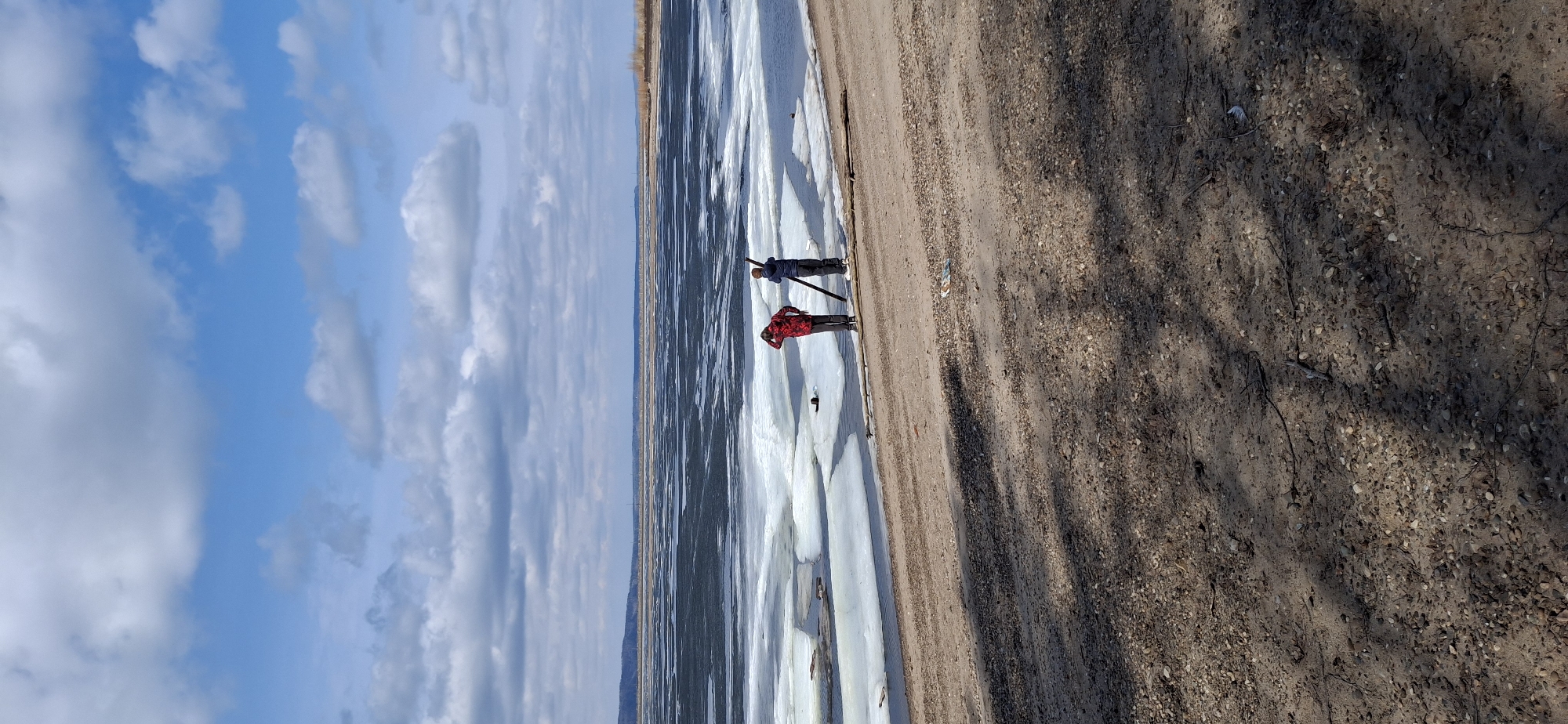 An outdoor scene captures two individuals standing at the edge of a partially frozen body of water in Chaikovsky, Russia, likely during a cold season such as winter or early spring. The sky is a vivid blue, dotted with numerous white, fluffy clouds.
Two people are the primary subjects. One person, closer to the viewer, wears a red and black plaid jacket and dark pants, and appears to be looking out across the water. The second person, positioned slightly behind and to the left, is dressed in a dark blue or black jacket and pants, and holds a long, dark stick or pole which extends towards the icy surface. They both seem to be observing the conditions of the water and ice.
The body of water is extensively covered with large, fragmented sheets of ice, varying in thickness and texture, with darker areas indicating patches of open water between the ice floes. This suggests either a recent freeze or thaw. In the far distance, a faint line of land or horizon is visible, possibly covered with sparse, dry vegetation.
The immediate foreground features a sandy-gravelly shore, speckled with small pebbles and fragments of twigs. Dark, elongated shadows stretch across the ground, cast by unseen objects, hinting at bright sunshine and a clear day, possibly late morning or early afternoon. No visible text is present in the image.