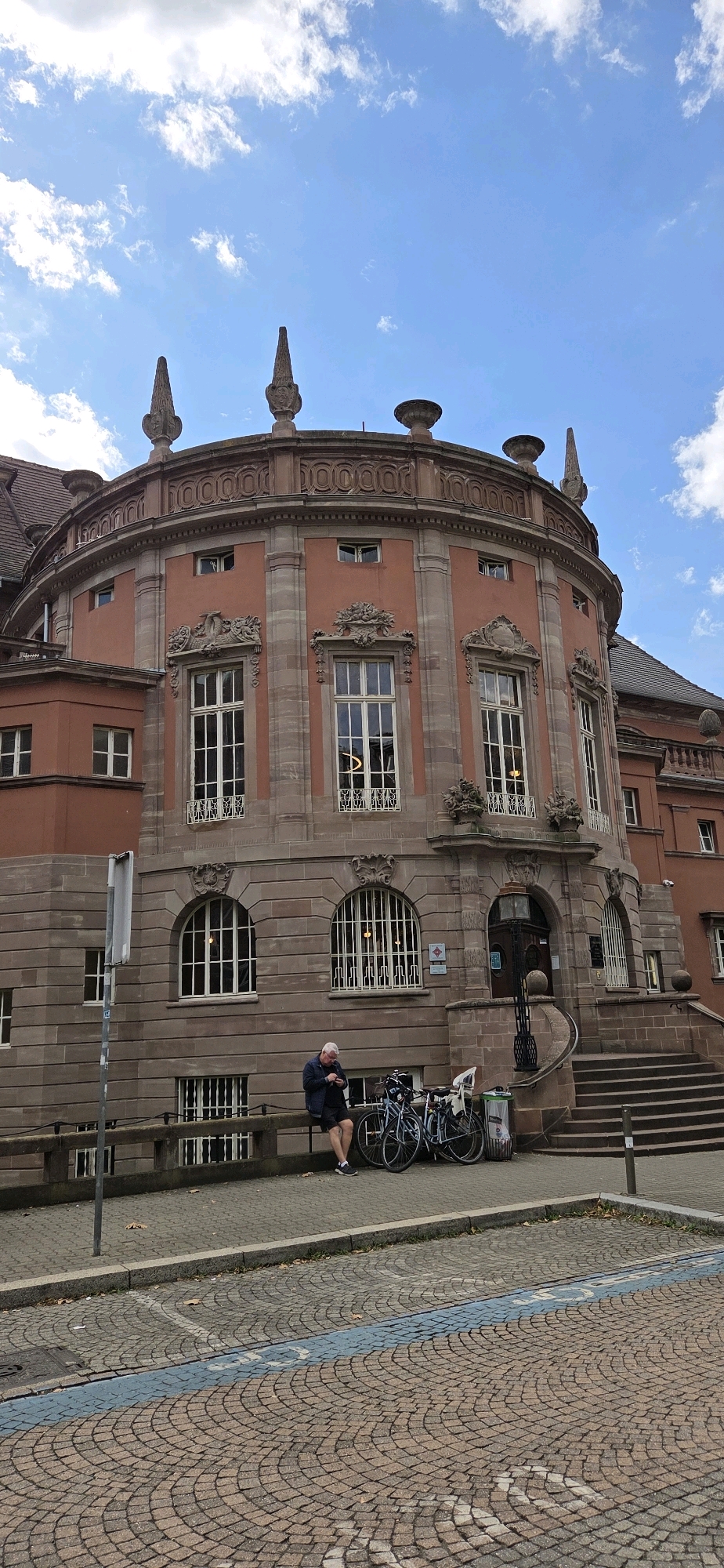 The image shows a round, reddish-orange building with stone accents, likely an administrative or cultural building, in Strasbourg, France. The building features arched windows with decorative grates and ornate carvings above them. On the top of the building, there are decorative spires and urns. In front of the building, a man is sitting on a low wall, looking at his phone. Two bicycles are parked next to him. The ground in front of the building is paved with cobblestones, and there is a blue line painted on the ground indicating a parking area. The sky above is blue with scattered white clouds. The overall scene suggests a quiet, daytime setting in an urban environment.