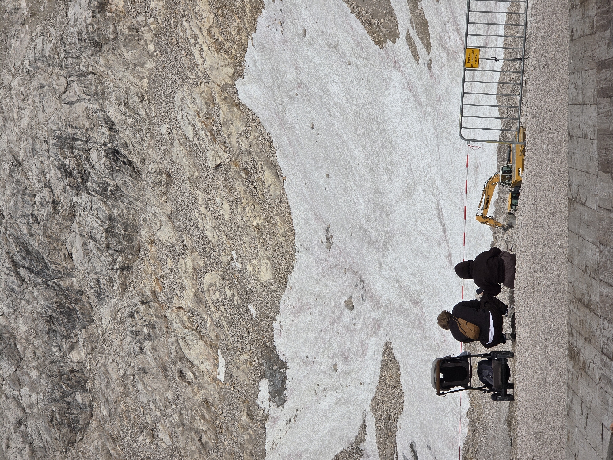 The image depicts a mountainous landscape with patches of snow. In the foreground, two individuals are seated on the ground, facing away from the camera, with a baby stroller beside them. A yellow excavator is partially visible behind a metal barrier in the middle ground, and a yellow sign with German text is attached to the barrier. The rocky, mountainous terrain suggests a high-altitude location. This scene is likely located in the Bavarian Alps near Garmisch-Partenkirchen, Germany, possibly a construction or maintenance site on a mountain.