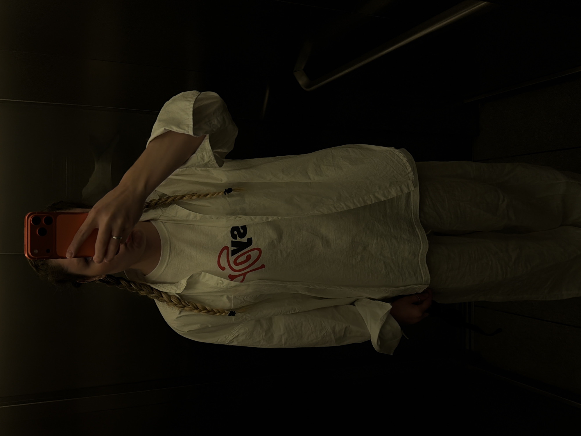 A woman is taking a mirror selfie in what appears to be an elevator. She's holding an orange smartphone with her left hand, and her face is partially obscured by the phone. She is wearing a white t-shirt with a large red logo, and a loose-fitting white button-down shirt. Her hair is styled in a long braid. The elevator interior is dark and minimally furnished, with what looks like a grab bar on the wall. The location is not identifiable as Tel-Aviv based on the visual content.