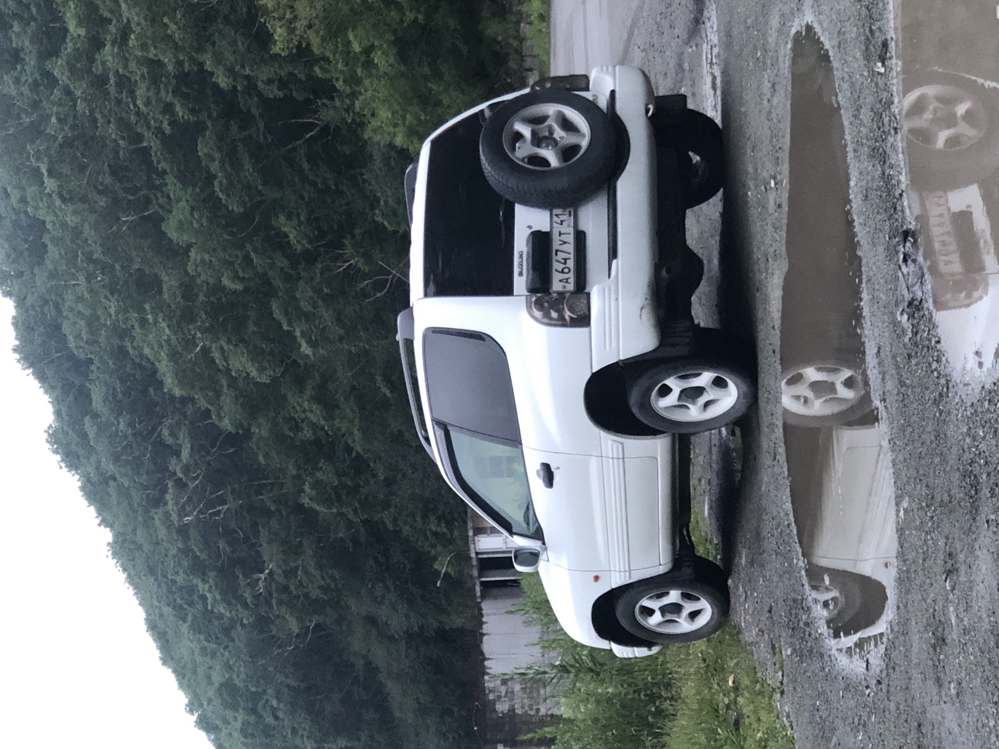 A white Suzuki Vitara is parked on a gravel road next to a puddle reflecting the vehicle and the surrounding environment. The car is facing right, with its rear slightly angled towards the camera. The spare tire is mounted on the back of the car, and a license plate that reads "A647YT 41" is visible. In the background, a dense, green forest covers a hillside. To the left of the car, there is a weathered, abandoned building. The sky is overcast, contributing to the muted lighting of the scene. The image was taken in the city of Petropavlovsk-Kamchatsky, Russia, likely in a rural or undeveloped area given the natural landscape and the presence of the abandoned structure. There are no people visible in the image.