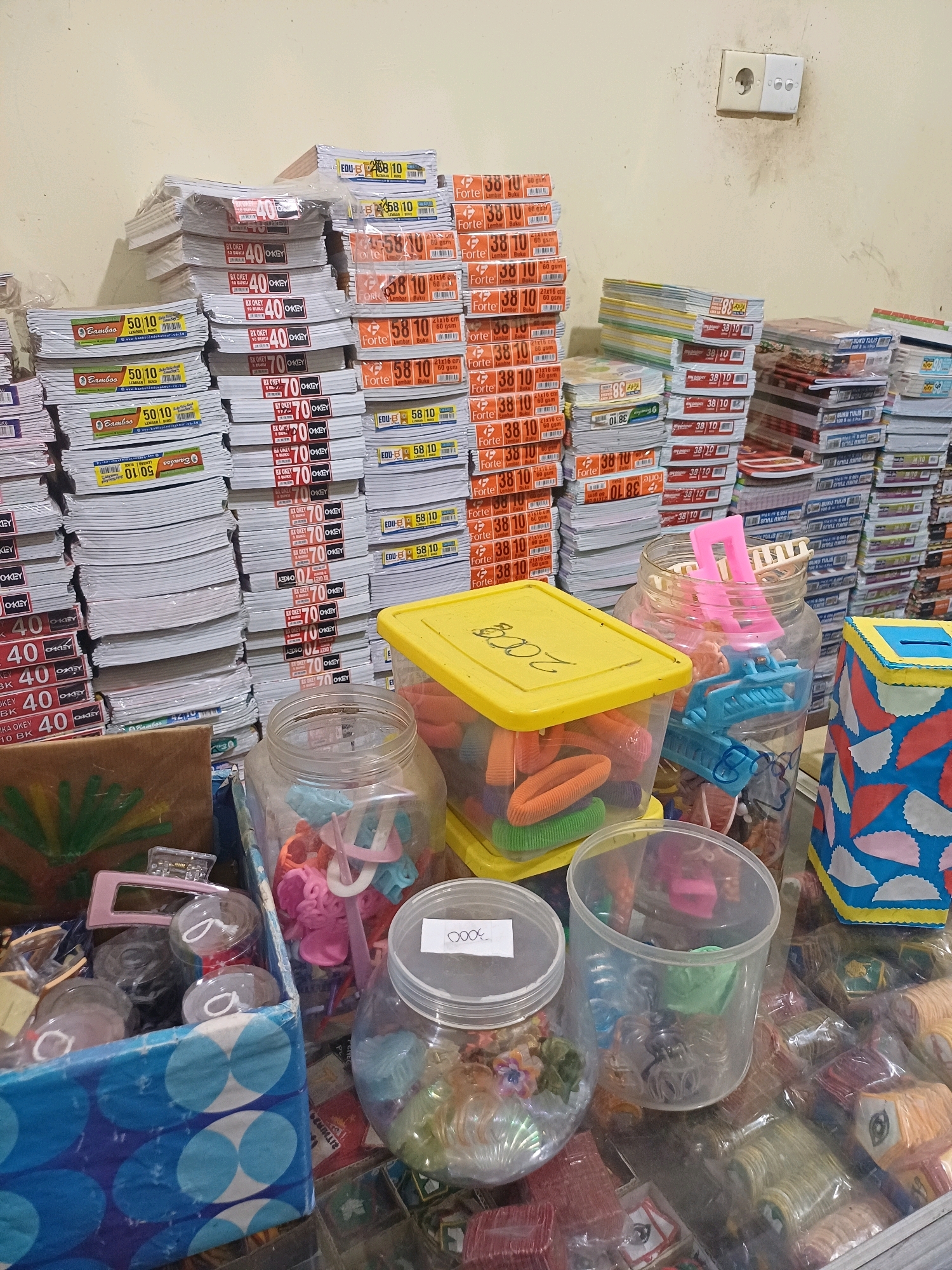 The image depicts a retail setting, likely a small shop or stall, within the city of Dumai, Indonesia. The primary focus is on stacks of notebooks and stationery items. Numerous notebooks, varying in size and branding (such as "Bamboo," "OKEY," and "Forte"), are neatly piled high, suggesting a supply of school or office supplies. In the foreground, several plastic containers hold an assortment of small, colorful items. These include hair accessories like hair clips and scrunchies, as well as other miscellaneous small objects. One container has a label that appears to read "2000," possibly indicating a price. The background shows a light-colored wall with a simple electrical outlet, a common feature in Indonesian buildings. The lighting is even, suggesting an indoor environment, possibly illuminated by fluorescent or natural light from an unseen source. The overall impression is one of a well-stocked stationery store.