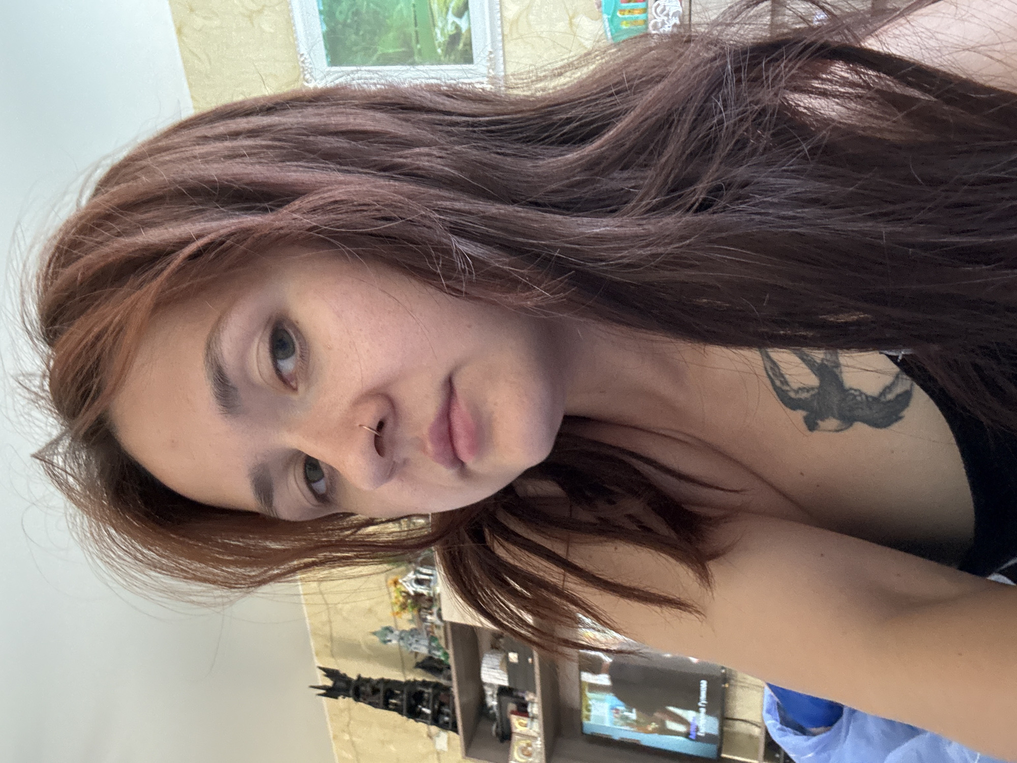 A young woman with long, dark auburn hair and a nose ring is looking towards the camera. She is wearing a dark tank top, and a black tattoo of a bird is visible on her chest. The background appears to be an indoor setting, possibly a bedroom or living room, with shelves containing various decorative items, a television displaying a program, and framed pictures on the wall. The overall lighting suggests it is daytime. The image does not provide specific information about the location within the city of Naberezhnye Chelny, Russia.