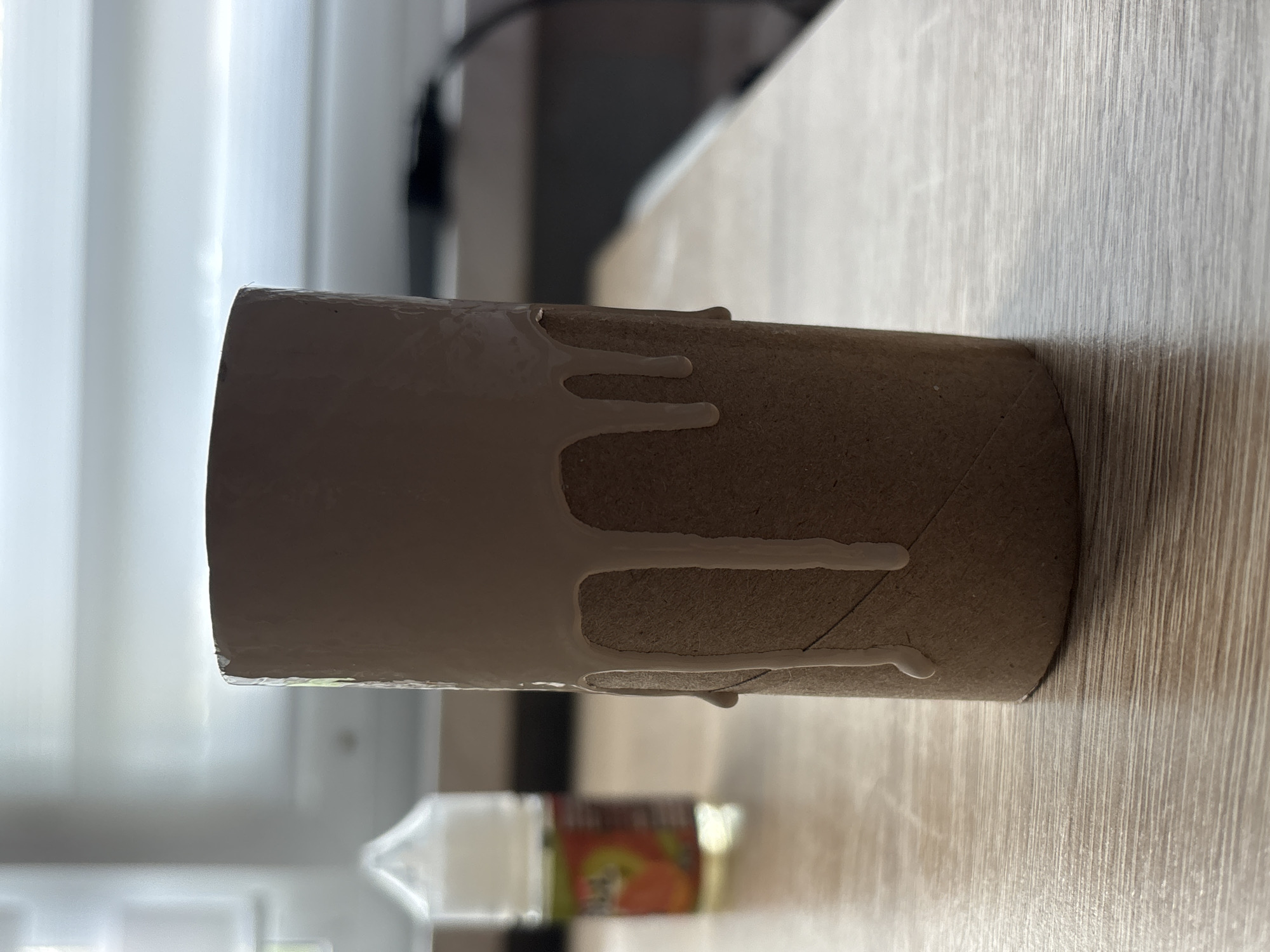 The image shows a close-up, eye-level shot of a cardboard toilet paper tube standing upright on a light-colored wooden surface. The top portion of the tube is coated with a thick, white substance that is dripping down the sides in several places, creating a melted or painted effect. In the background, out of focus, is a clear plastic bottle with a white cap, containing a yellow liquid and a label with orange and green elements. Further in the background, a window with horizontal blinds is visible, letting in natural light. The scene appears to be indoors, possibly on a table near a window. There are no people visible in the image. The overall impression is a still life arrangement with an artistic or experimental element. The location in Saint Petersburg, Russia is not evident from the image itself.