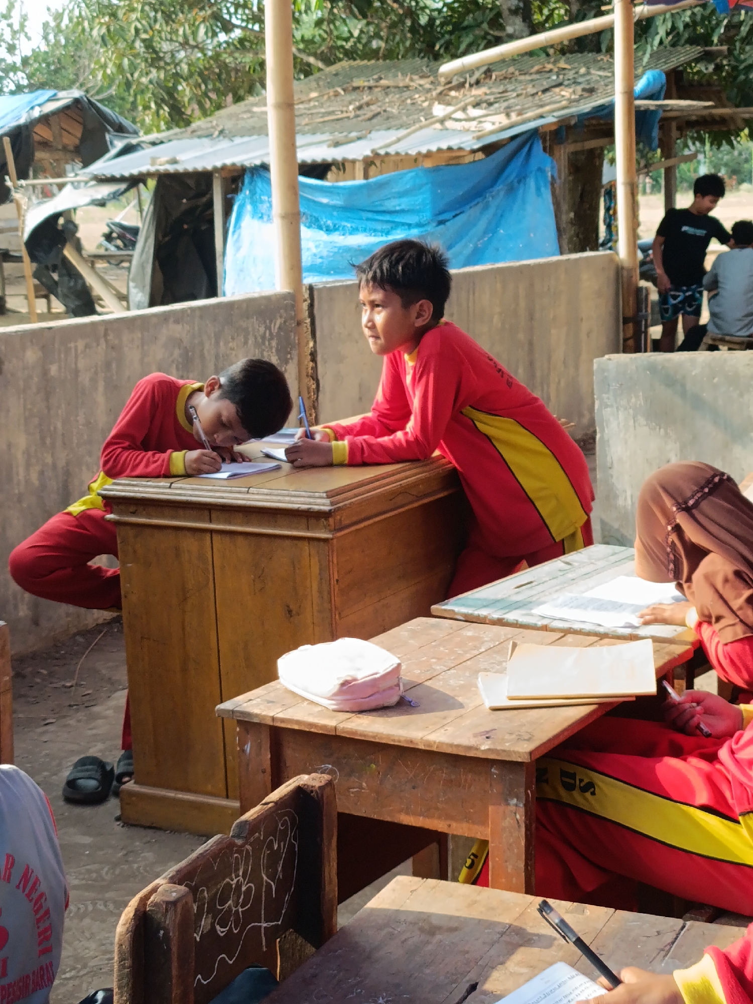 In Sumberejo, Indonesia, a classroom scene unfolds outdoors. Two young boys, dressed in red and yellow school uniforms, are seated at wooden desks. One boy leans over his desk, writing on a piece of paper with a blue pen. The other boy, to his right, also wears a similar uniform and is engaged in writing as well. Behind them, a third student, wearing a brown hijab and a red uniform, is also seated at a desk, looking down at their work. The environment appears somewhat rustic, with a makeshift roof structure in the background supported by bamboo poles, and corrugated metal roofing on adjacent structures. Other students and adults are visible in the distance, suggesting a school setting. The ground is dirt.