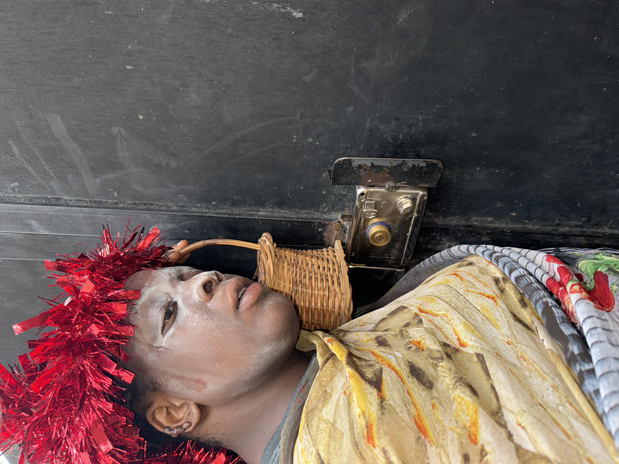 A person lies on their back wearing a tinsel headpiece and a patterned shirt, with a small woven basket positioned near their neck. The person is under a dark surface with a metal lock mechanism attached. This scene is located in Sabon Tasha, Nigeria.