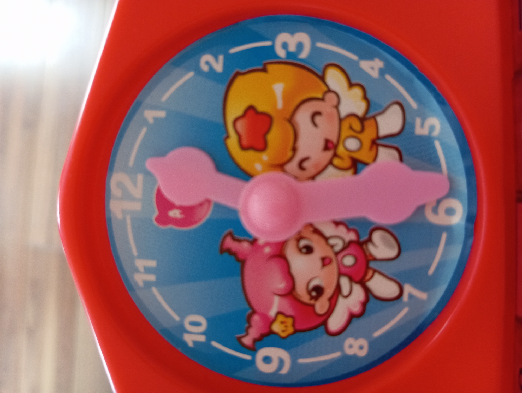 A close-up, eye-level, overhead shot shows a red plastic toy clock with a blue face. The clock has cartoon characters with pink and yellow hair, wings, and star accents, positioned at the 9 and 3 o'clock positions. A pink plastic clock hand is positioned between the 11 and 12 o'clock positions, indicating the time is approaching 12. The background appears to be a wooden surface. There is no discernible information to indicate this image is located in Hlynytsi, Ukraine.