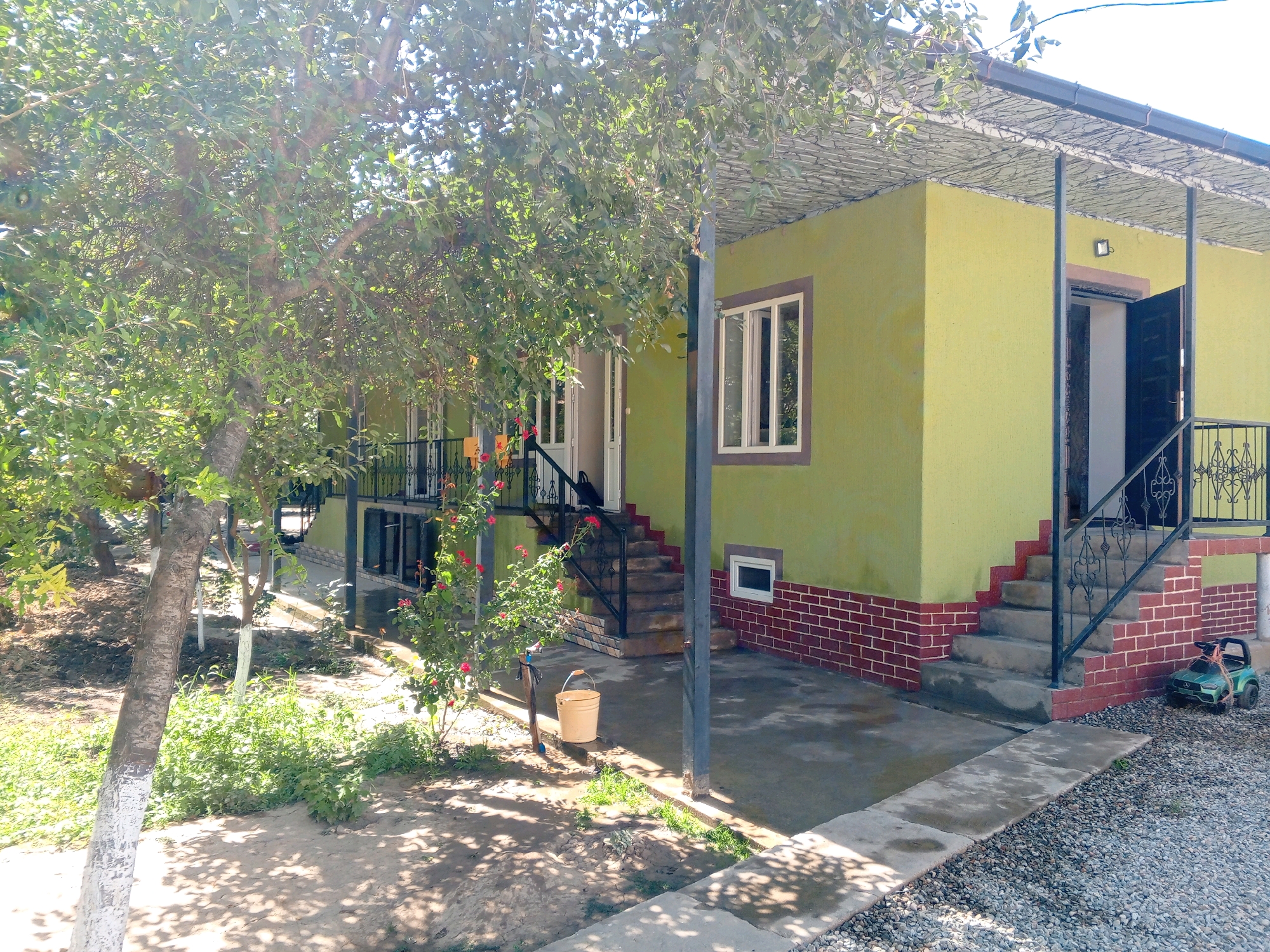 A single-story, lime-green building with a brick-patterned base is situated in a yard with trees and vegetation. The building has a covered porch supported by pillars, with a corrugated roof. A set of concrete stairs with a black metal railing leads up to a dark-colored door. To the left of the stairs, there is a window with white trim. Another window, also with white trim, is visible on the side of the building. A small rose bush with red flowers is planted near the stairs. In the yard, there is a light-colored bucket, a hose faucet, and a small green toy car. The ground in front of the building is paved with concrete, transitioning to gravel on the right. The scene is bathed in sunlight, casting shadows from the trees. The location is in the city of Kofarnihon, Tajikistan.