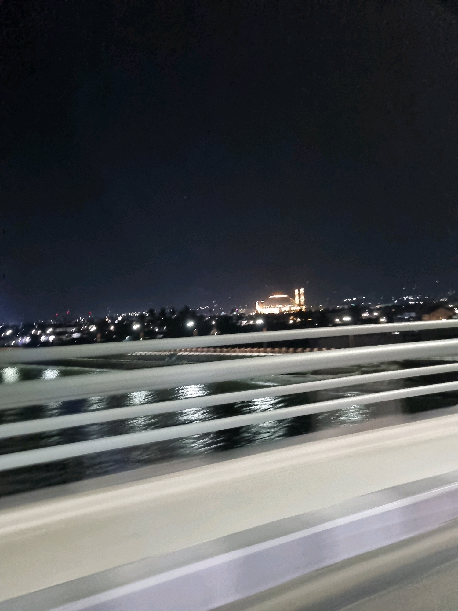 This image shows a nighttime view of the city of Balaroa, Indonesia, from a bridge. The sky is dark with a few scattered lights visible in the distance. A large, illuminated building with a distinctive dome and two towers is prominent in the center of the frame. The foreground is dominated by the white railing of the bridge, with reflective surfaces below showing the lights of the city. The overall impression is a cityscape at night, with a focus on the illuminated architecture.