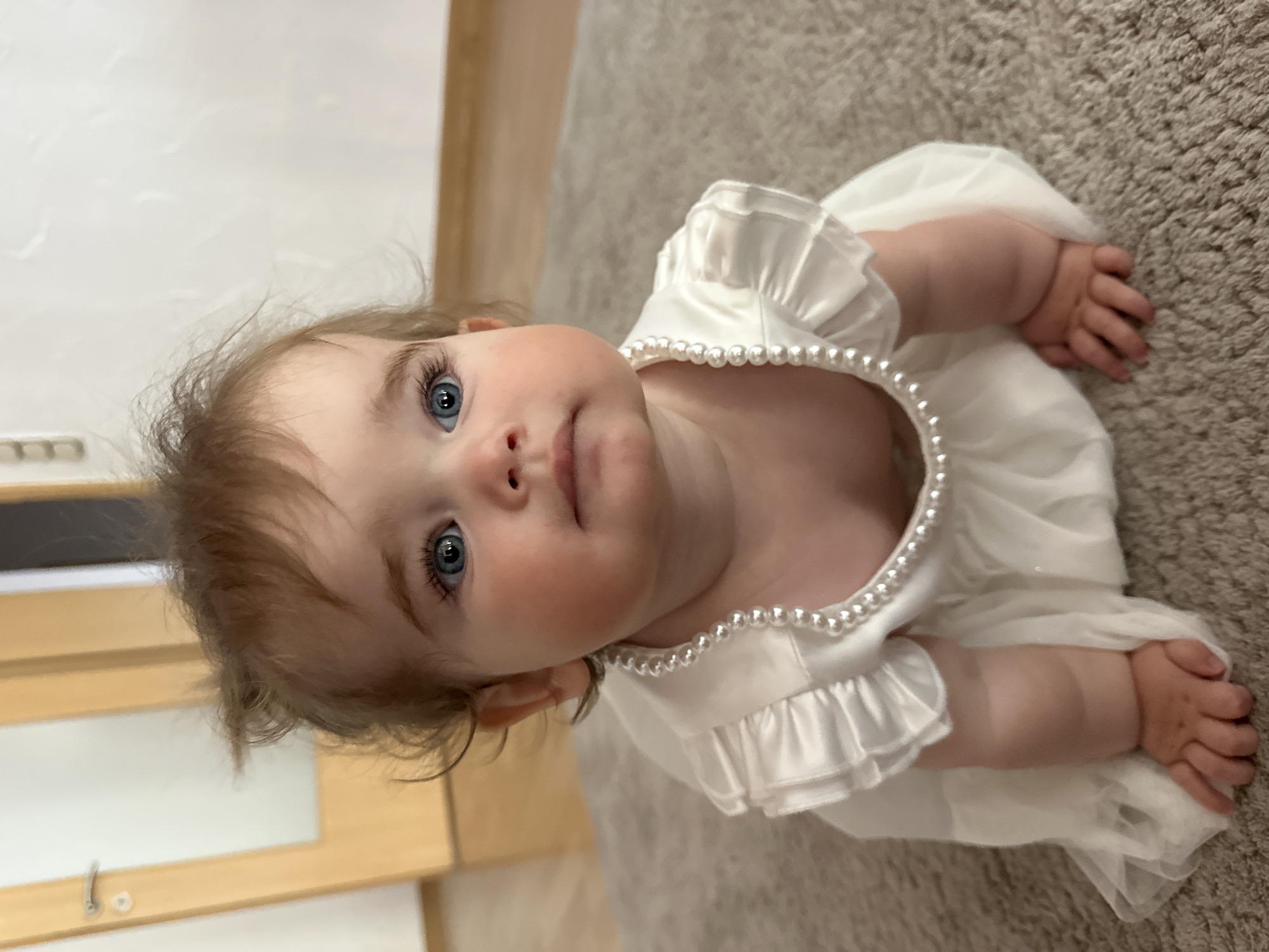 A baby wearing a white dress with a pearl necklace is lying on a beige carpet. The baby is looking up at the camera with blue eyes. The location is inside a building, but Niederquembach, Germany, is not identifiable from the image.