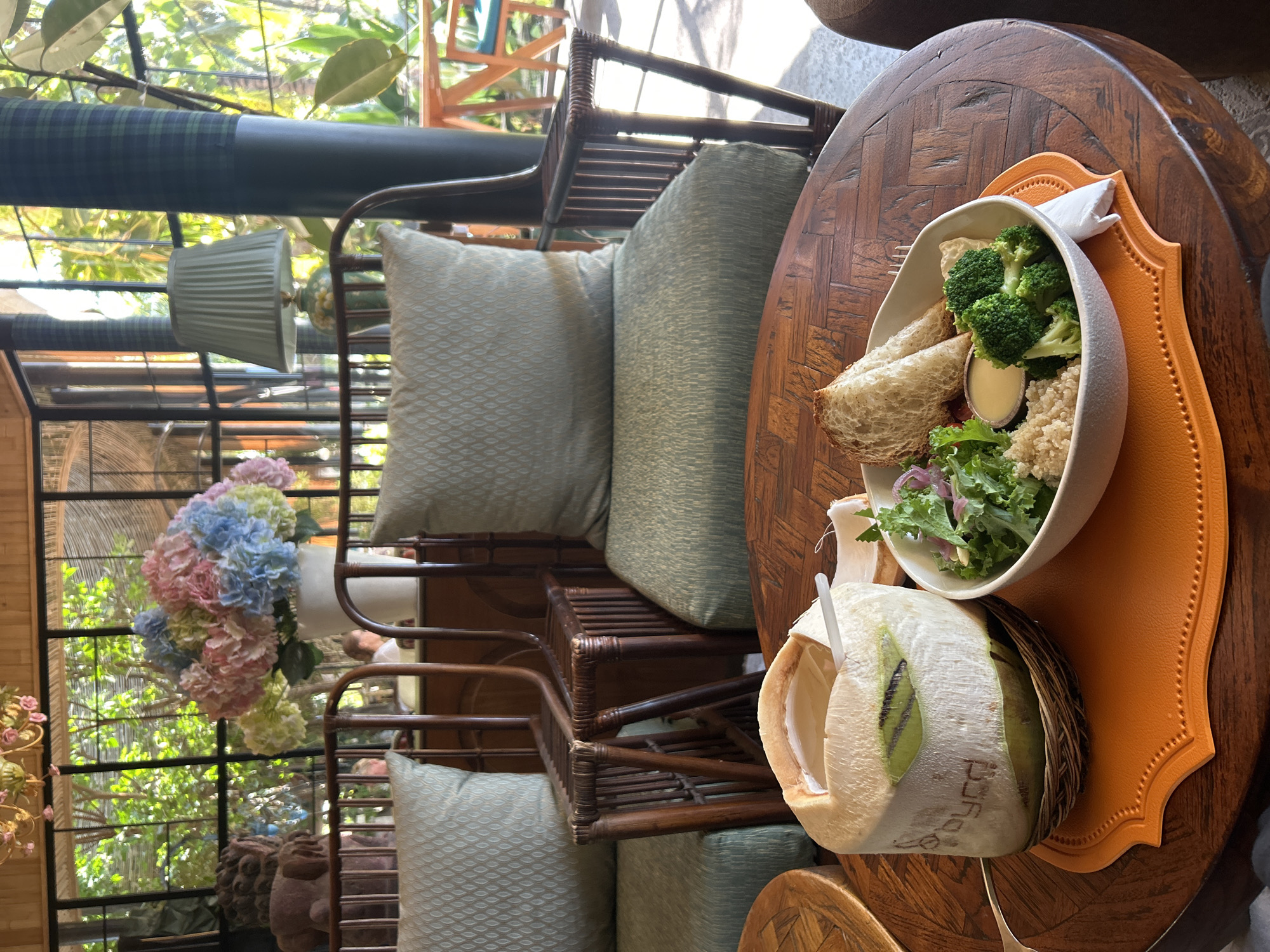 A half-cut coconut with a straw rests on a woven coaster on a wooden table. A bowl containing quinoa, salad greens, broccoli, and a small dish of dressing sits to the right of the coconut. Two slices of toasted bread are placed in the bowl. The background shows two wicker chairs with green patterned cushions, a large vase of colorful hydrangeas, and a window overlooking lush greenery. A lamp with a pale green shade is visible in the background. The scene appears to be set in an outdoor cafe or restaurant, possibly in Denpasar, Indonesia, given the tropical foliage. There are no visible people actively engaged in the scene, but blurred figures can be seen in the background.