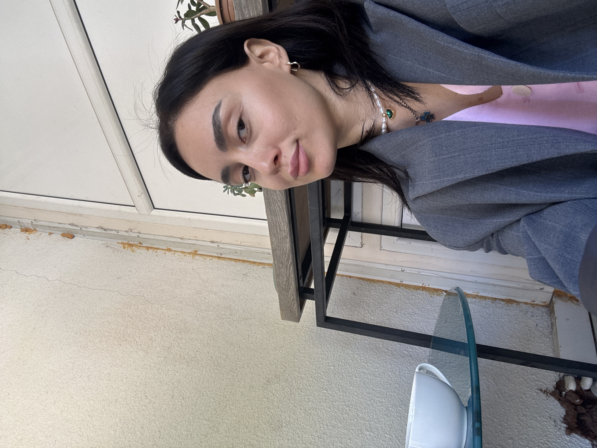 A medium shot captures a woman in what appears to be an outdoor setting. She is wearing a gray blazer over a light pink top and jewelry including a pearl necklace and earrings. She is positioned near a glass-topped table. Behind her is a wall, a door or window frame, and a potted plant. While the photograph does not contain enough information to definitively confirm the location in Tbilisi, Georgia, the outdoor setting with building elements is consistent with urban environments.