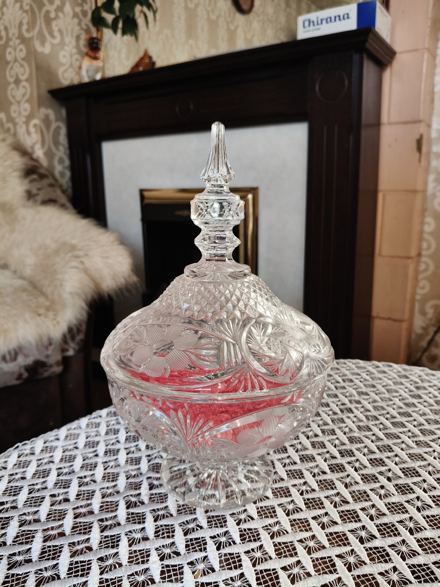 A detailed, high-angle medium shot shows a crystal candy dish filled with red candy sitting on a white crocheted doily. The dish, with a pointed lid, has intricate cut-glass detailing. Behind the dish, a dark wooden fireplace mantel is visible, with a gold-framed mirror above it. A white fuzzy blanket is draped over a chair to the left of the mantel. A small white box with blue text that reads "Chirana" is on top of the mantel to the right. The background wallpaper is a light beige with a subtle damask pattern. The scene appears to be indoors, likely in a living room or dining room.
