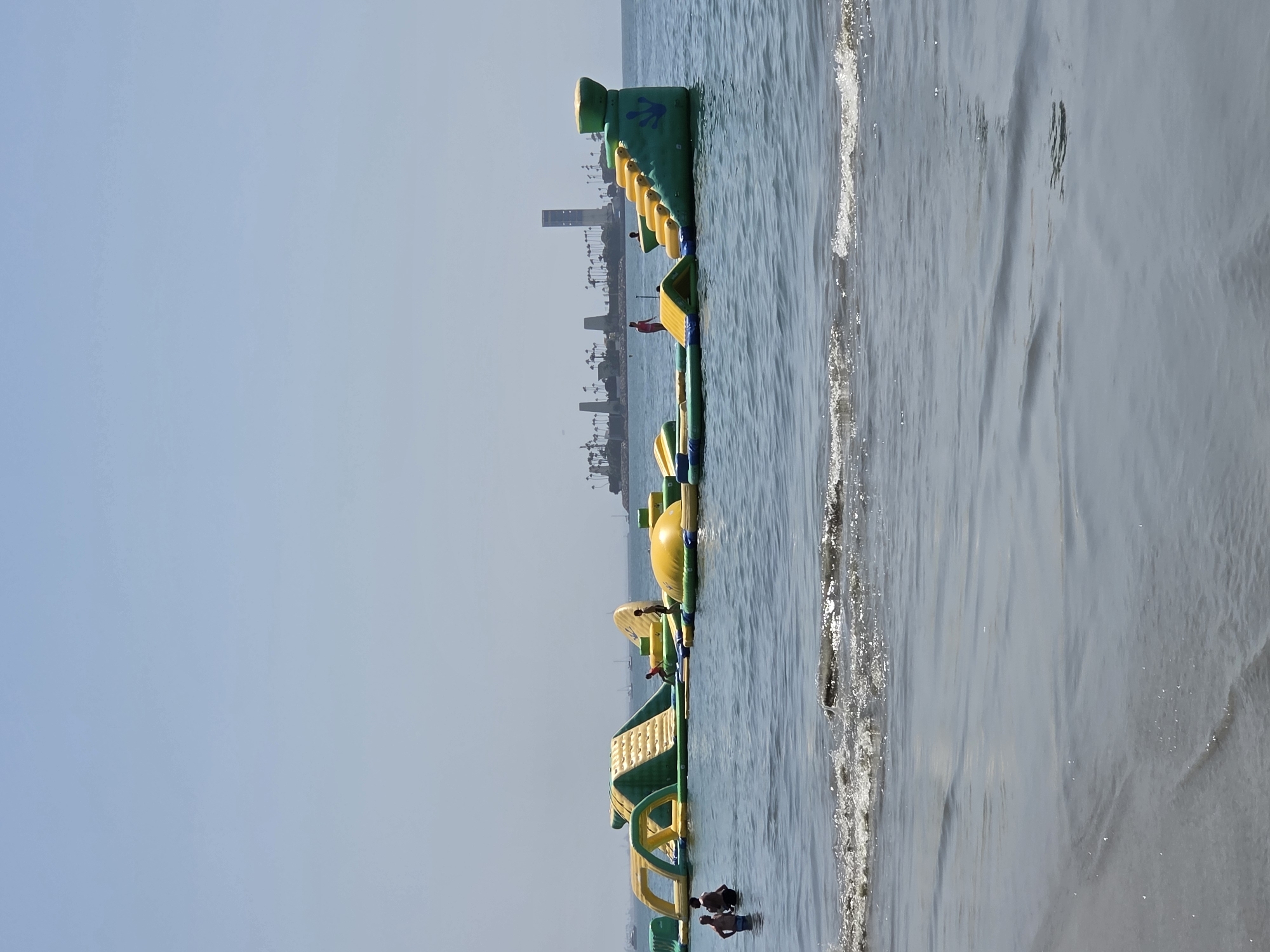 The image shows an inflatable water park on the ocean in Long Beach, California. The water park is composed of several yellow and green inflatable structures, including slides, climbing walls, and obstacle courses. There are a few people in the water, some on the inflatable structures and some wading in the shallow water near the shore. In the background, there is a cityscape with buildings and palm trees, and several boats on the water. The sky is clear and blue, and the sun is shining.