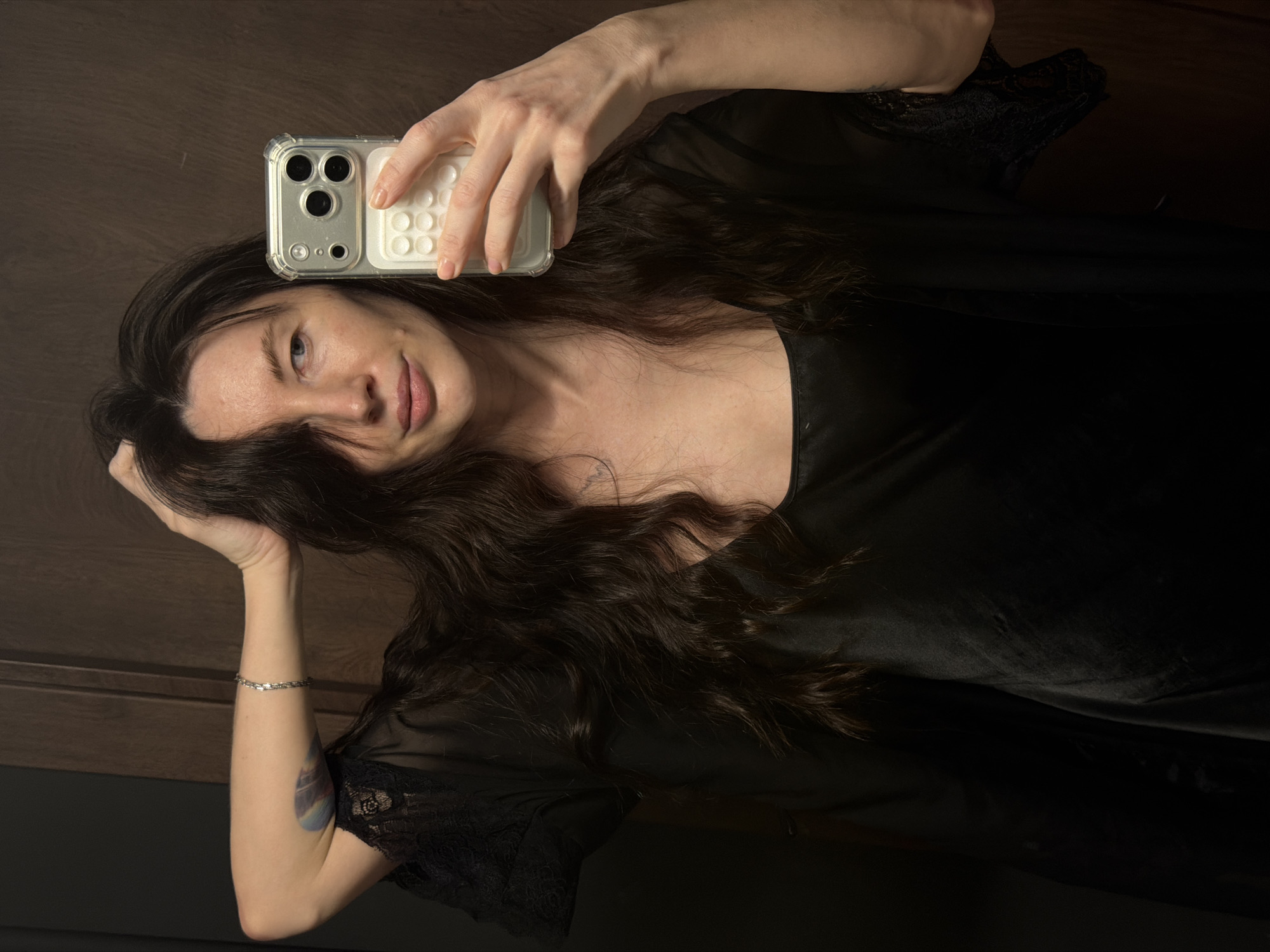 A woman is taking a selfie indoors. She has long, dark hair that partially covers her face and is wearing a black garment with lace trim. She is holding a smartphone in her right hand with a clear case with a white, tactile back. She is wearing a silver bracelet on her left wrist, and a small tattoo is visible on her left forearm. The background appears to be a wooden door or wall. The location is unknown and there is no mention of the city of Perm, Russia.