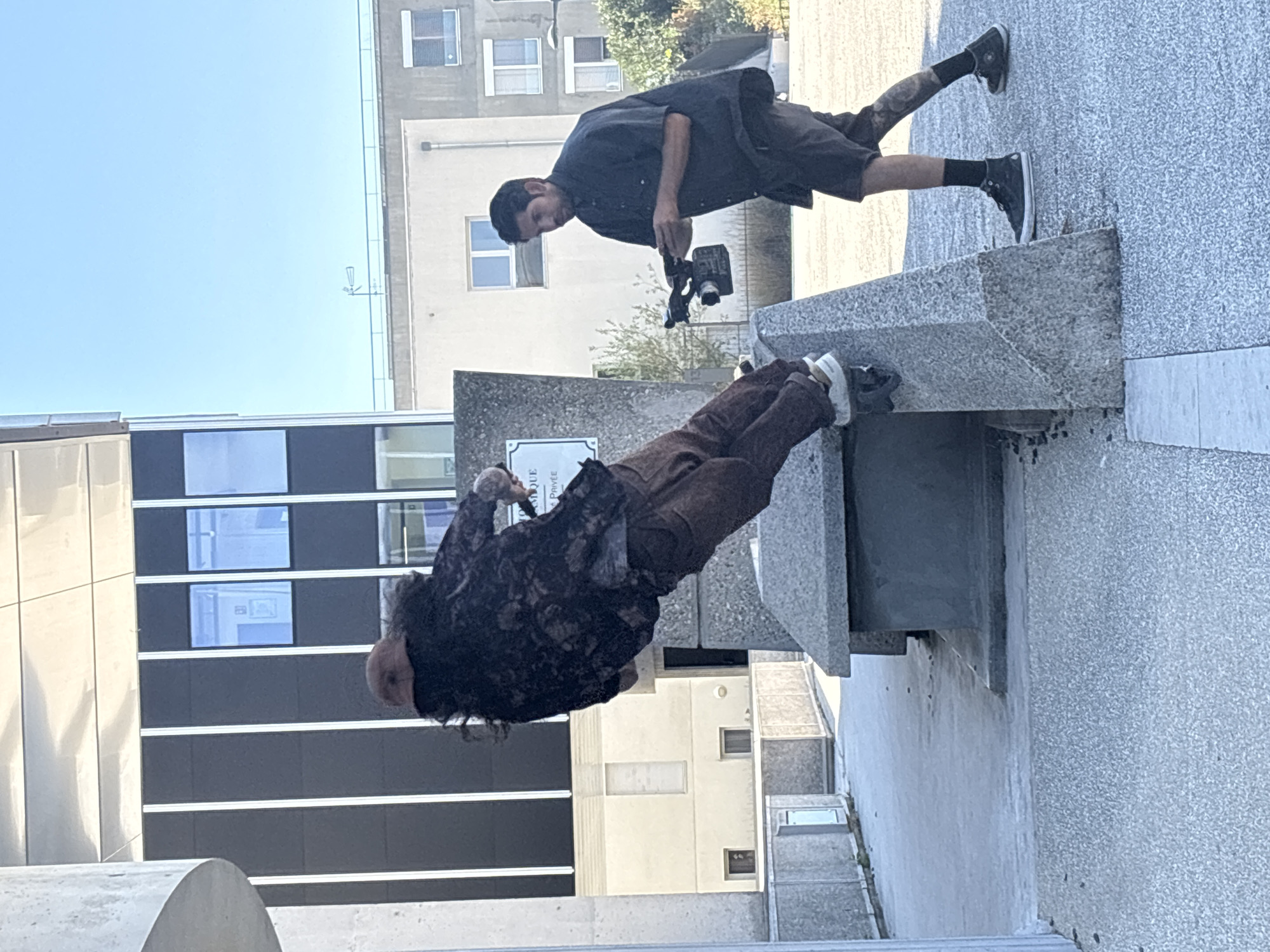 A man is captured mid-action, leaning precariously over a concrete structure, his body angled downwards as if in the midst of a parkour move or stunt. He is wearing brown pants, a dark, patterned jacket, and light-colored sneakers. His long, dark hair is flung out behind him, indicating rapid movement. To his right, another man stands attentively, holding a video camera and appearing to film the action. This second man is dressed in a dark short-sleeved shirt, dark shorts, black socks, and black sneakers. The scene unfolds outdoors on a bright, sunny day, with modern urban architecture forming the backdrop. The ground is paved with a textured material. This location is likely in Lyon, France, given the context provided.