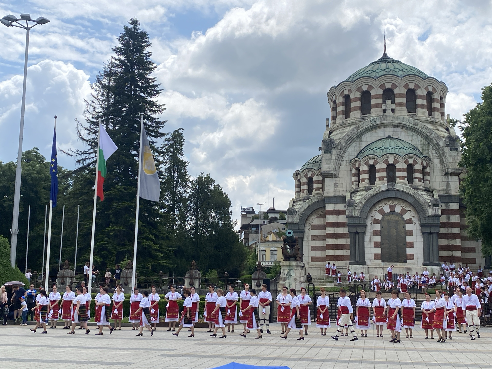 The image depicts a group of people in traditional Bulgarian folk costumes performing in a public square. The performers are wearing white shirts, red embroidered skirts, and black shoes, and they appear to be part of a choreographed dance. In the background stands the St. George the Victorious Chapel Mausoleum, a historical landmark located in Pleven, Bulgaria. Flags, including the European Union and Bulgarian national flags, are visible to the left. The sky overhead is cloudy.