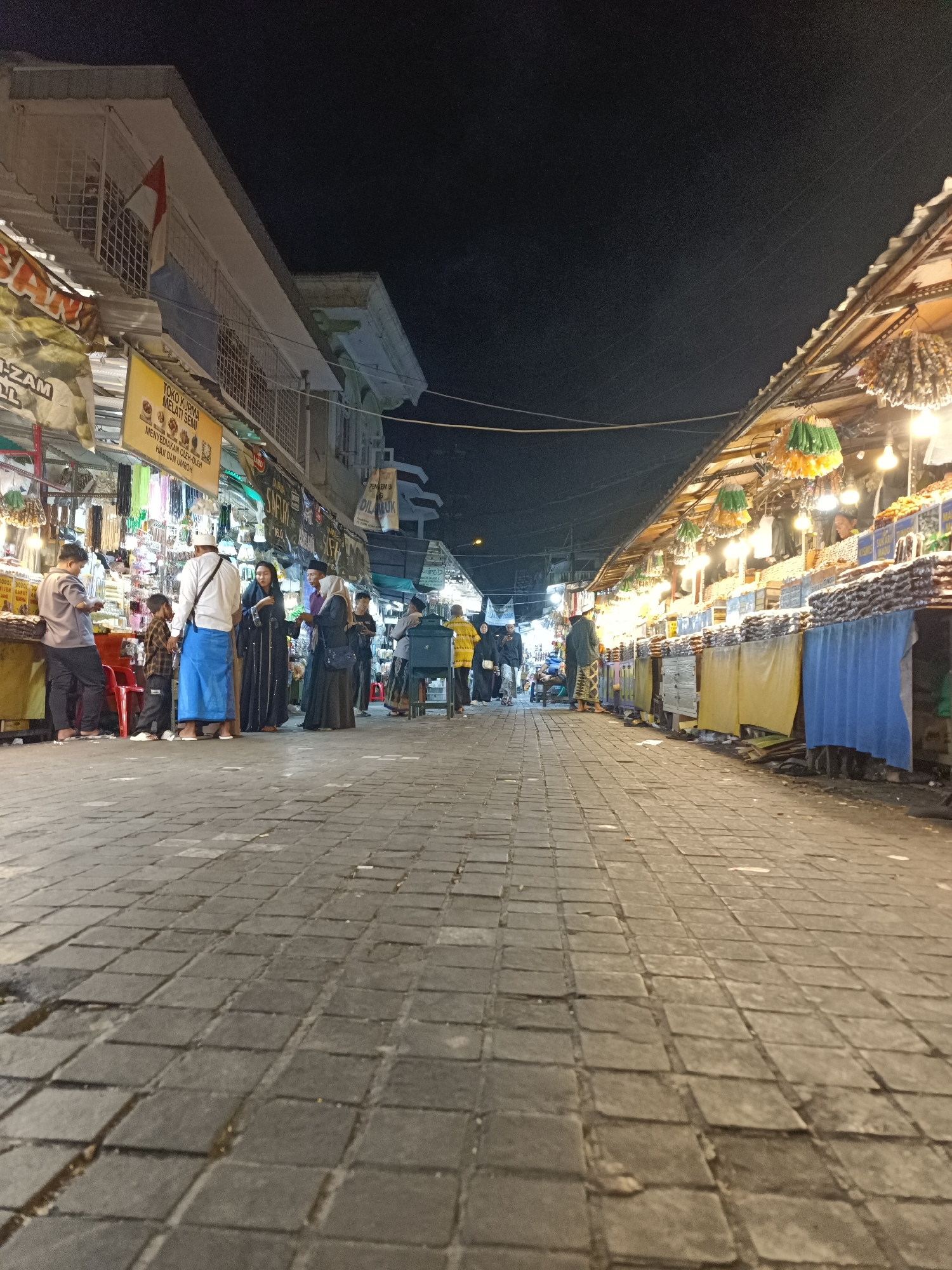 The image depicts a bustling night market scene in Surabaya, Indonesia, likely near a religious site given the attire of some individuals and the presence of religious merchandise. The perspective is low, looking up from a paved walkway made of square stones. On the left, market stalls display various goods, including what appear to be necklaces or prayer beads, and banners with Indonesian text such as "TOKO KURMA MELATI SEMI". Several people are browsing or standing in front of these stalls.
On the right, another row of market stalls is visible, illuminated by bright lights, showcasing a variety of products, possibly food items or other commodities, arranged in piles and within containers. Some stalls have colorful fabric coverings, like blue and yellow. The architecture of the buildings behind the stalls suggests a mix of traditional and modern construction, with some showing balconies and ornate details. A red and white flag, possibly the Indonesian flag, is seen on the building to the left. The overall atmosphere is lively, with people engaged in shopping and socializing under the night sky.