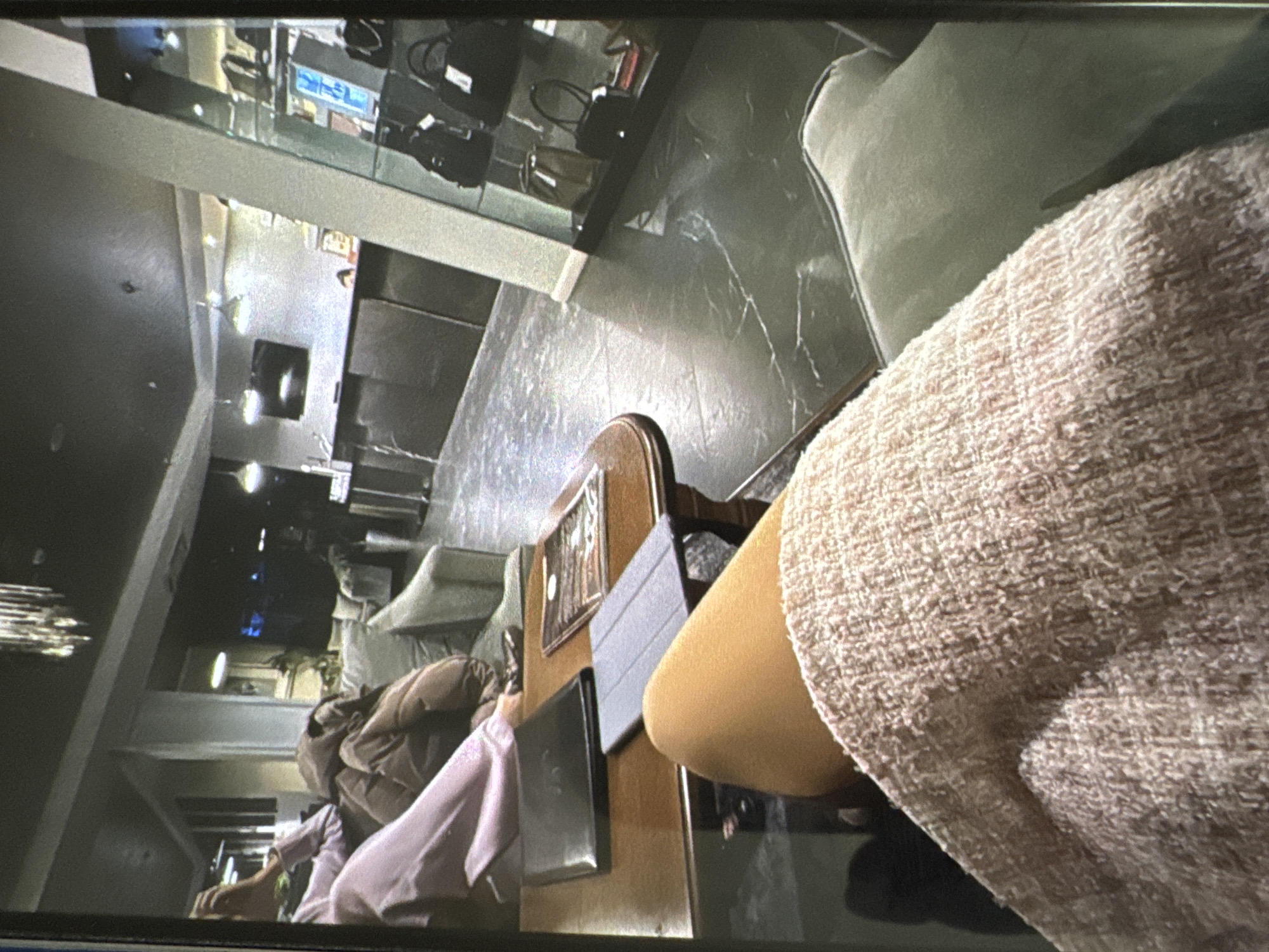 The image depicts an indoor lounge or waiting area, possibly within a hotel or business establishment. In the immediate foreground, the perspective is from a seated individual, showing their upper right thigh covered by a light-colored, textured, possibly tweed, garment, and a bare leg (thigh/knee area) beneath. To the right, a segment of a light green or grey upholstered cushion is visible.
In the midground, a wooden coffee table with a light brown finish occupies the central space. On the table, a black rectangular object (possibly a folder or tablet case), a closed grey tablet cover, and a magazine or brochure are arranged. Behind the table, two individuals are seated on a dark-colored sofa. The person on the left wears a light purple long-sleeved top; their left arm is raised, and a hand is near their head. The person on the right is clad in a brown, puffy jacket or similar outerwear. Further back on the left, another lighter-colored sofa is visible.
The background features a dark ceiling from which a multi-tiered chandelier hangs on the left. To the right, a dark, reception-style counter or desk spans the width of the area. Behind this counter, a person's head is faintly visible, suggesting an attendant. Above the counter, two pendant light fixtures and a wall-mounted television screen are present. On the far right, a tall glass display case showcases multiple black handbags and accessories on several shelves. The floor throughout the visible space is dark grey or black, exhibiting white, marbled veining. The walls are predominantly light grey or off-white.
The specific location within the city of Vratsa, Bulgaria, cannot be forensically determined from the visual information provided in the image.