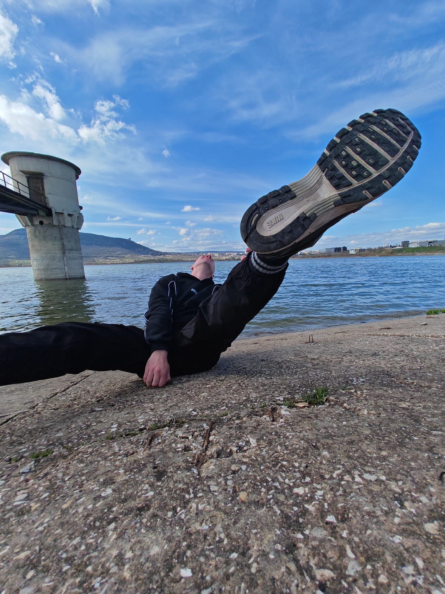 A person is lying on a concrete surface beside a body of water, extending one leg upwards so that the sole of their athletic shoe is facing the sky. The person is wearing black pants and a black hooded sweatshirt with white stripes on the cuffs. They appear to be looking up at the shoe. The water is calm, and in the background, there is a concrete structure in the water, appearing to be some kind of tower or pump house connected to the shore by a bridge. In the distance, a hill or mountain is visible. Based on available geographical information, this scene is located at Yazovir Ticha Reservoir, outside the city of Shumen, Bulgaria.