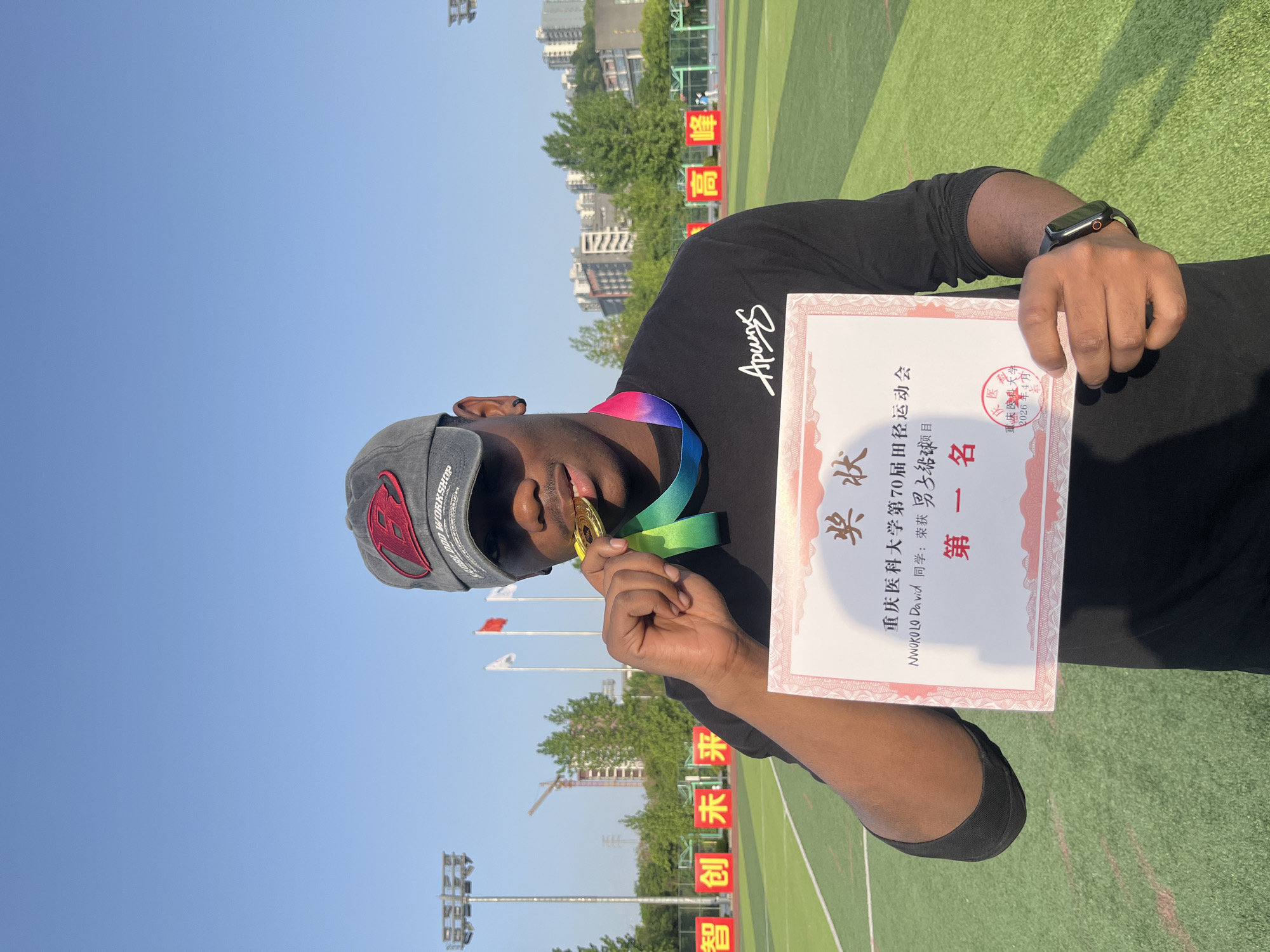 The image shows a man holding a gold medal and a certificate on an athletic field. The man is wearing a black t-shirt and a gray baseball cap. The certificate is written in Chinese and English and states that NWOKOLO David has won first place in the men's shot put event at the 70th Chongqing Medical University Games. The background shows a green athletic field, flags, and buildings in the distance. The text "智创未来“ is also visible. These signs suggest the location is in Chongqing High-tech Zone, China.
