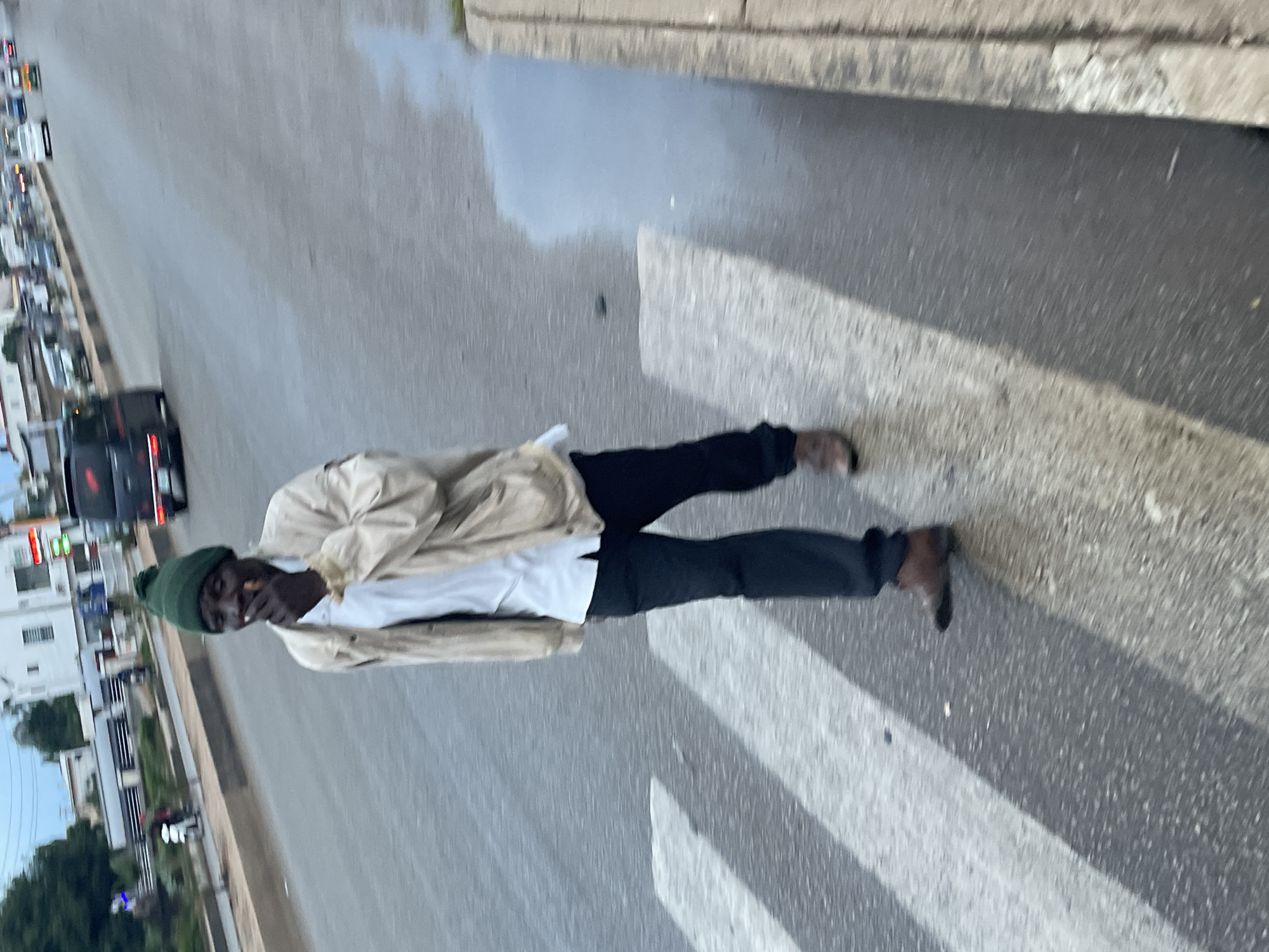 A man in a green beanie, a light-colored jacket over a white shirt, and dark pants is walking across a crosswalk. He is holding a cigarette to his mouth. The crosswalk is wet, as is a portion of the road to the right, suggesting recent rain. In the background, traffic is visible on a multi-lane road, with cars and a black van. Buildings and trees line the side of the road. The scene appears to be in Gura Top, Nigeria, during daylight hours, with an overcast sky.