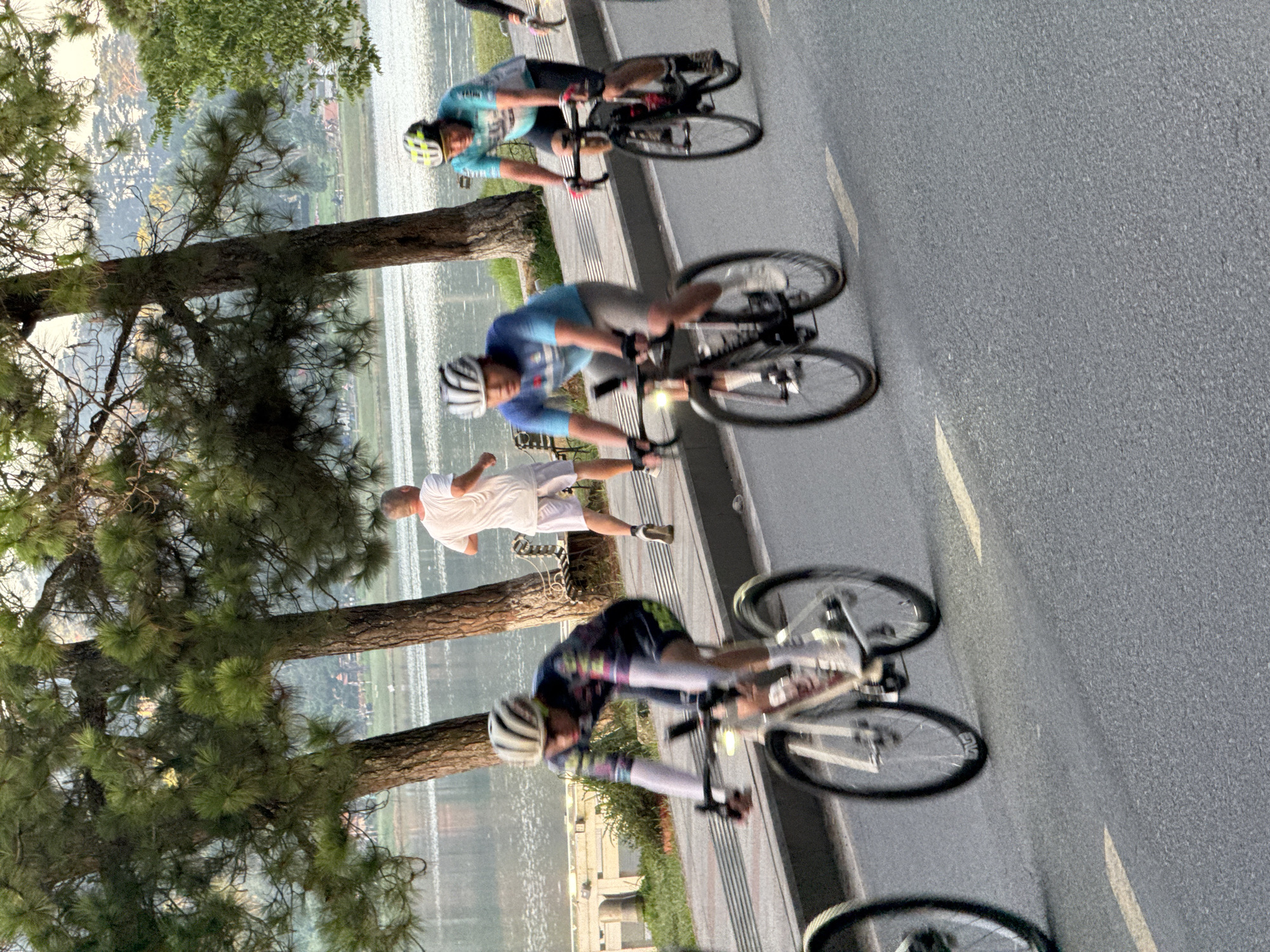 A group of cyclists are seen riding on a paved road in Da Lat, Vietnam. The scene takes place near Xuan Huong Lake, with the lake visible in the background and lined with trees and greenery. In the foreground, two cyclists in full racing gear are actively pedaling, their bikes in motion. One cyclist, in a colorful jersey, is on the left, while another in a blue jersey is to his right. Further back, a man in a white t-shirt and shorts is jogging. The image captures a moment of athletic activity in a scenic urban park environment.