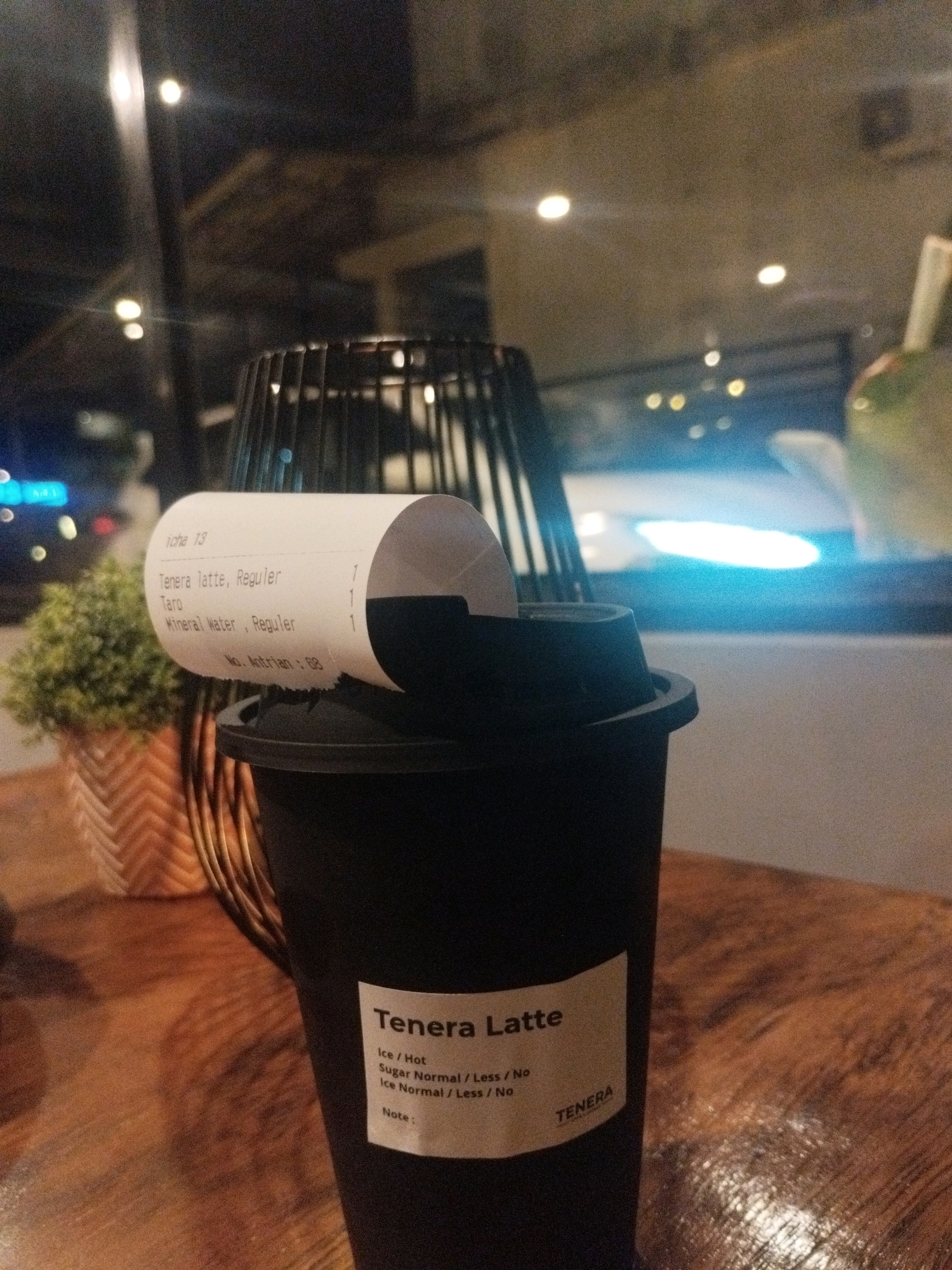 The image depicts a close-up, indoor scene featuring a black cup with a receipt partially unrolled on top of it. The receipt has text indicating an order for "Tenera latte, Reguler", "Taro", and "Mineral Water, Reguler". A small green plant in a patterned pot is visible to the left of the cup. The background shows a dimly lit environment with reflections of lights, suggesting it could be a cafe or restaurant. The overall setting appears to be a table surface, possibly made of wood. The location is identified as being in Sungailiat, Indonesia.