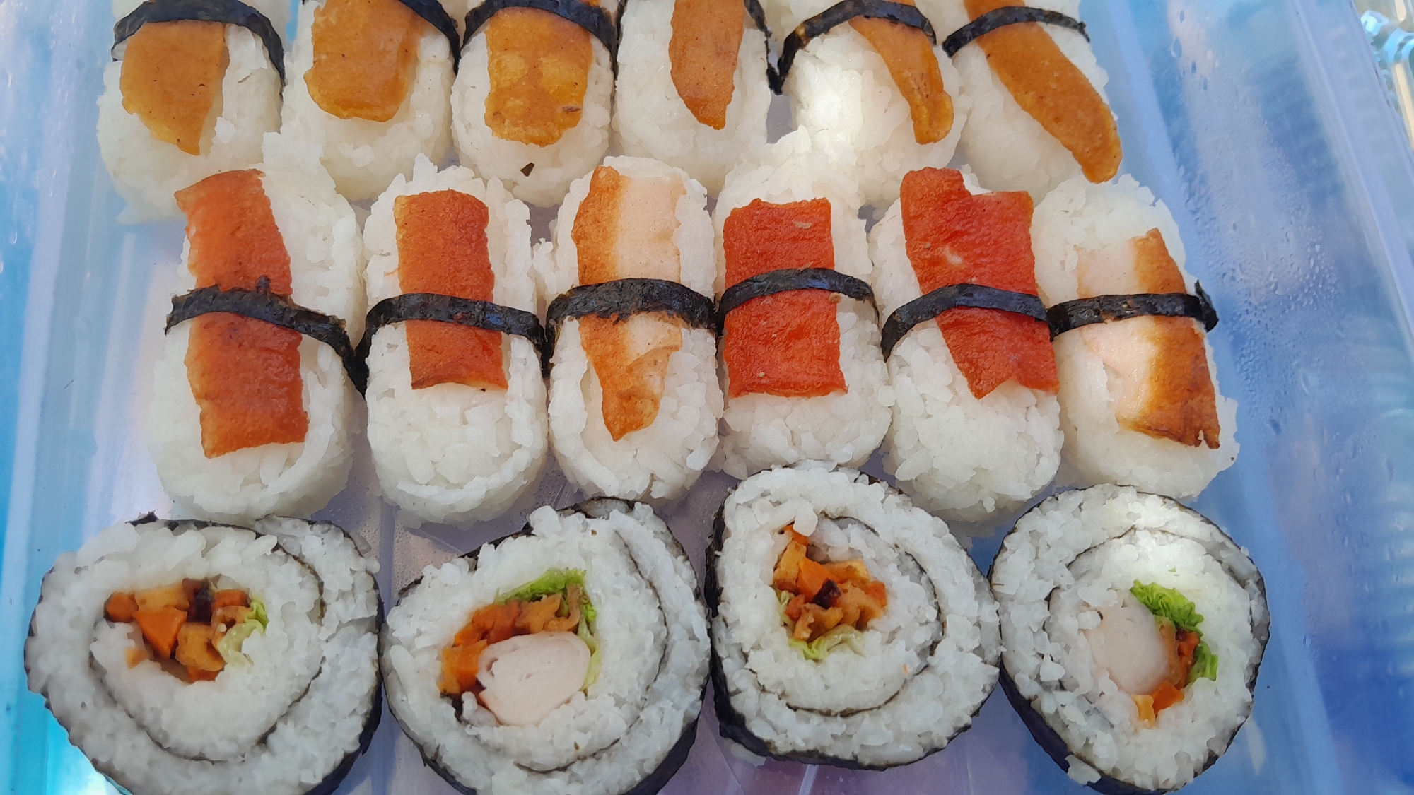 This is a top-down, close-up, eye-level shot of a variety of sushi rolls arranged in a clear plastic container. The sushi is the main subject, filling the entire frame. In the top row, there are four maki rolls with seaweed on the outside and rice, shredded carrots, lettuce, and a piece of white filling visible inside. In the middle row, there are six nigiri-style sushi pieces, each consisting of a ball of rice wrapped with a thin strip of nori (seaweed) and topped with a slice of what appears to be cooked meat or fish, some with a reddish-orange hue and others with a lighter brown hue. The bottom row contains six more nigiri-style sushi pieces, similar to the middle row, but the toppings are more consistently orange-brown, suggesting a different filling or preparation. The lighting is bright and overhead, casting slight shadows. The background is a blue, possibly plastic, surface with some condensation visible, indicating a cool environment. The image does not contain any people or specific landmarks that would indicate a location in Bandung City, Indonesia.