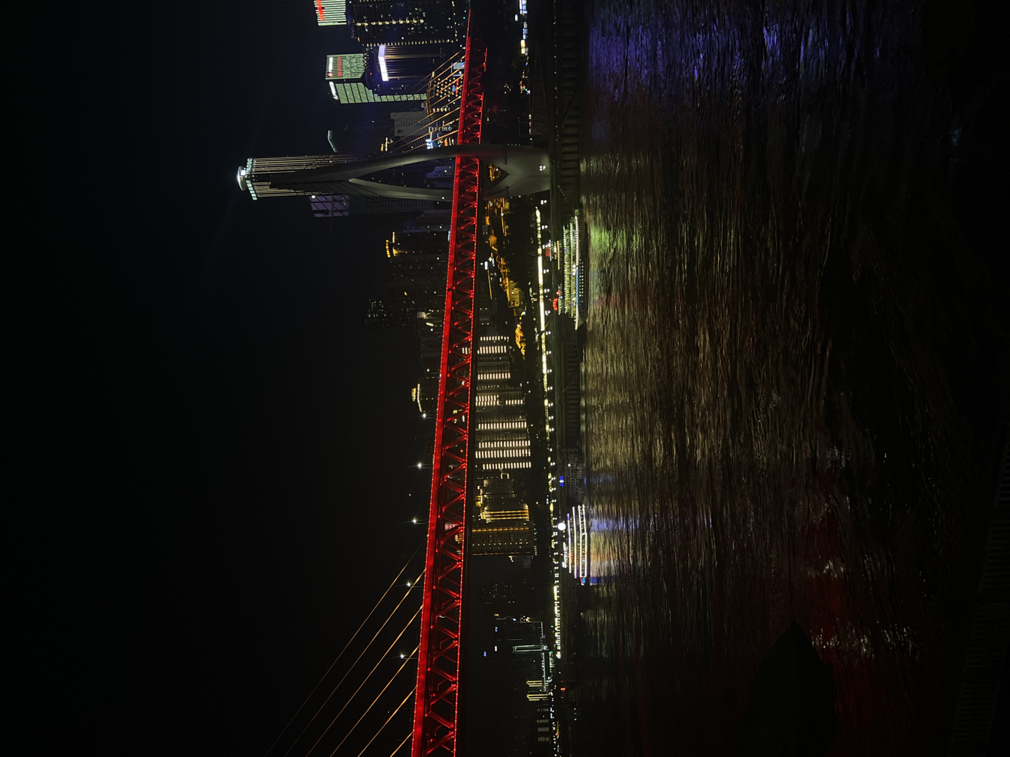 The image depicts a nighttime view of the Chongqing Yangtze River Cableway in Nan'an District, China. A bright red illuminated bridge spans across a dark body of water, reflecting the vibrant lights. Two illuminated tour boats are visible on the water, one closer to the foreground with multi-colored lights and another further in the background. The cityscape beyond the bridge is illuminated with numerous buildings, including a prominent skyscraper with vertical lighting. The overall scene is characterized by the contrast between the darkness of the night and the numerous artificial lights from the bridge, boats, and city.
