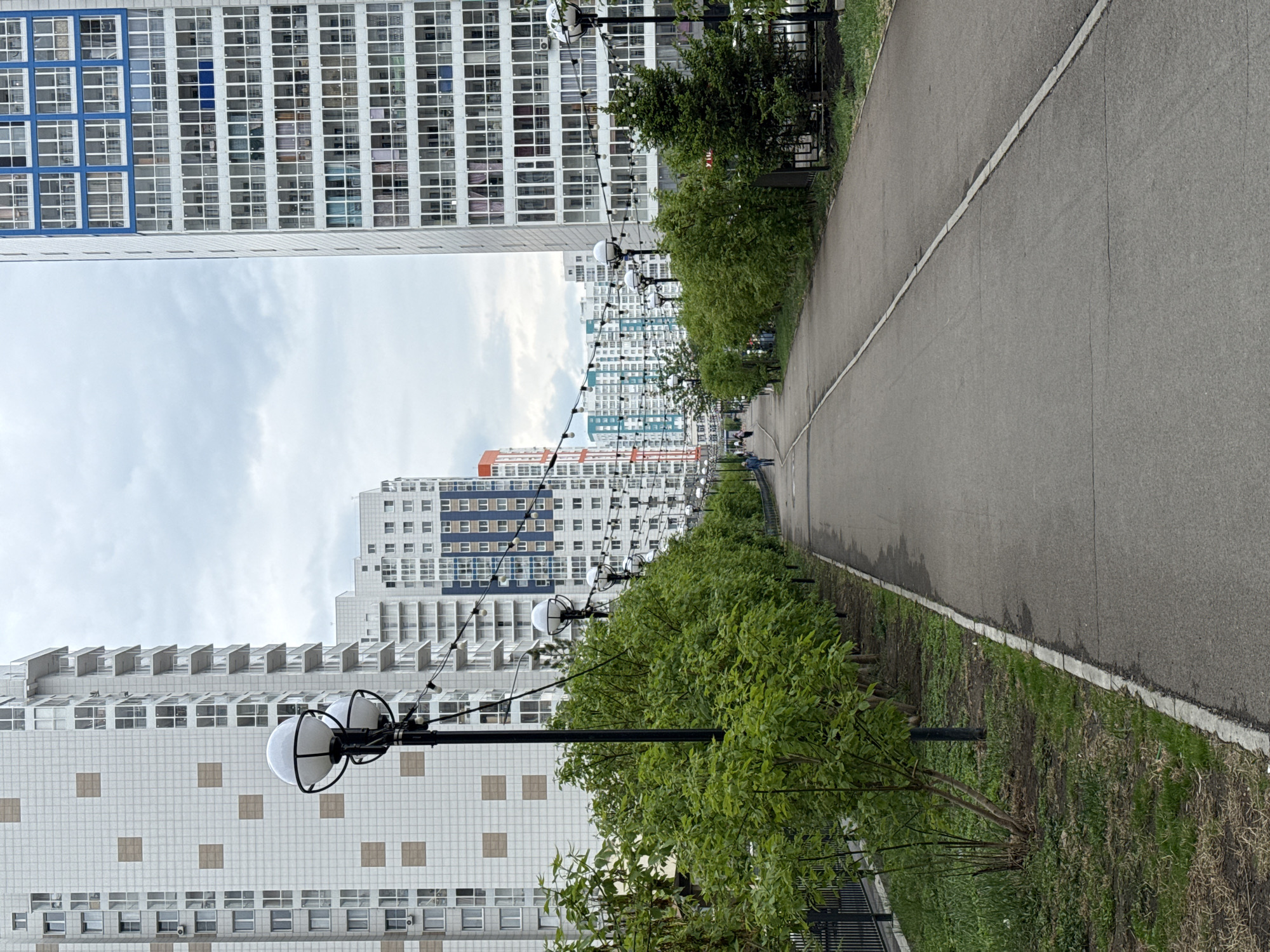 A gray asphalt road stretches between lines of modern high-rise apartment buildings in what appears to be an urban landscape. The buildings are primarily white and feature a grid-like pattern of windows, some with blue accents. Streetlights with globe-shaped fixtures line the path, interspersed with green shrubs and grass along the sides of the road. Two distant figures can be seen walking in the direction of the end of the road. The sky above is cloudy, suggesting overcast weather. Based on visual styles, this image captures a view along the "Park of Universiade" in Krasnoyarsk, Russia.