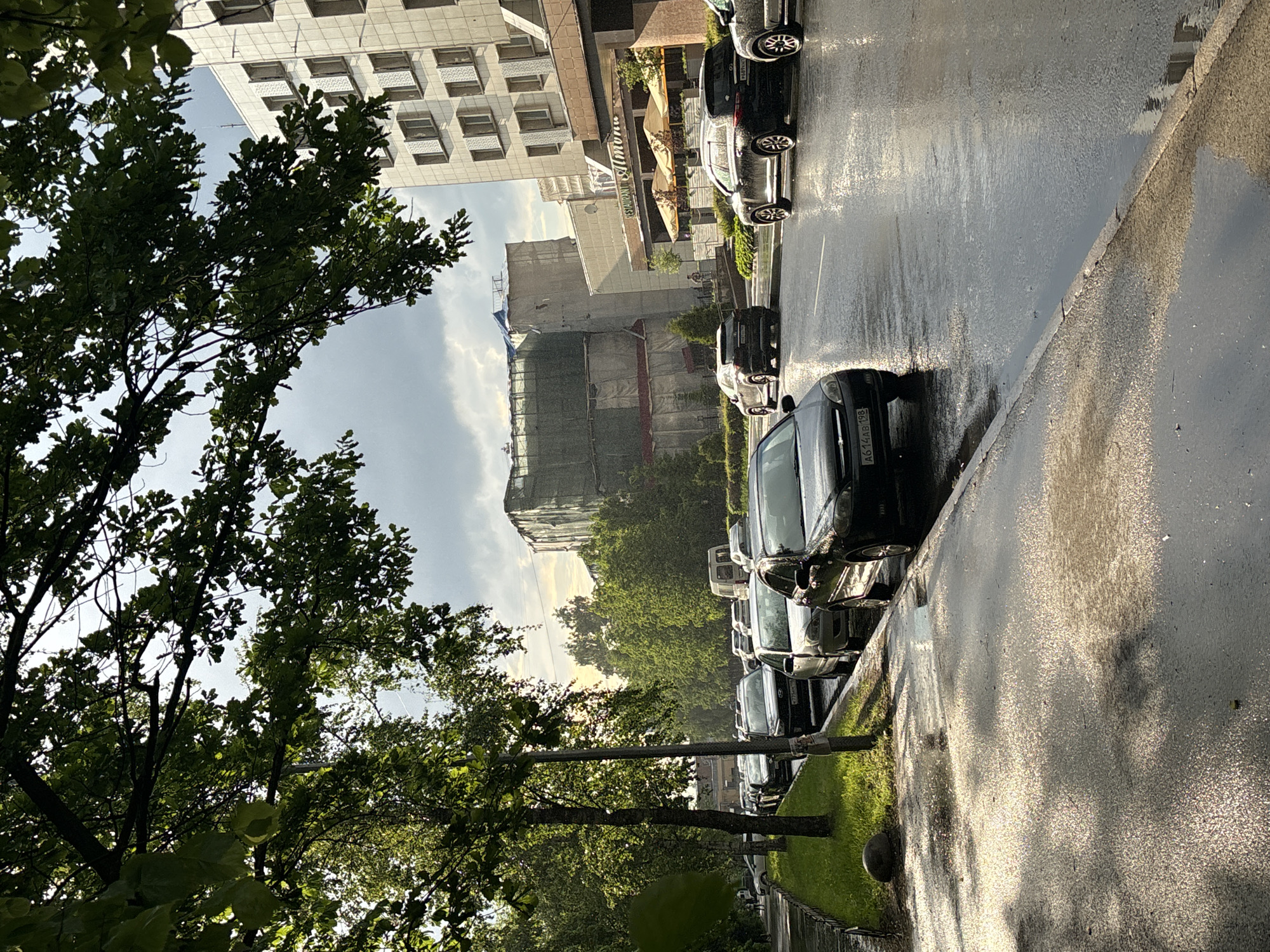 The image captures a wet urban street scene, viewed from a slightly elevated perspective, likely in a late afternoon or early evening setting due to the soft, reflective lighting. The street and sidewalk are visibly damp, indicating recent rainfall, with strong reflections of the sky and surrounding environment on the asphalt.
In the foreground and along the left side, lush green trees with dense foliage partially frame the view, their leaves appearing vibrant. A line of cars, mostly dark-colored sedans, are parked bumper-to-bumper along the right side of the street, facing away from the viewer. Visible license plates, such as "A614AB 198" and "P557XX 777", indicate the location is a Russian city, specifically Moscow (region code 198 and 777).
Beyond the parked vehicles, a narrow strip of green grass and more trees line the street. In the background, several buildings define the urban landscape. On the left, a tall, multi-story building features a light-colored facade with numerous rectangular or square windows arranged in a grid pattern. Towards the center-right, another significant building is undergoing construction or renovation, almost entirely enveloped in scaffolding and green protective netting. Between these structures, a restaurant awning is visible, bearing the text "RESTAURANT" and what appears to be "Al'mona" or similar.
The sky is partly cloudy, with brighter areas suggesting the position of the sun, which casts a gentle, diffuse light across the scene, enhancing the reflections on the wet surfaces. No people are discernible in the image, contributing to a tranquil, post-rain atmosphere in an active city environment.