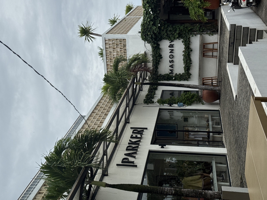This image depicts an outdoor commercial complex located in Uluwatu, Indonesia, under an overcast sky. The scene focuses on the ground level of a multi-story building featuring white exterior walls and an upper facade constructed with a grid pattern of light-colored louvers.
On the left, a storefront labeled "PARKER" with dark, embossed lettering is visible. Its large glass windows display various clothing items on racks and a mannequin in a light-yellow garment. An open-framed awning with a translucent roof extends over the "PARKER" entrance. To the right of "PARKER," another establishment features the name "MASONRY" in dark letters on a white wall, framed by dark green climbing vines. Below the "MASONRY" sign, a low, white, built-in bench is present, accompanied by two light-brown wooden bar stools. A large terracotta pot containing greenery stands near the entrance to the right of "PARKER," and another larger pot is partially visible on the far right.
The ground surface is paved with dark, interlocking bricks. A flight of light-gray concrete steps with white risers descends to a lower level on the right, where the front wheel and headlight of a white scooter are partially visible. Several palm trees with green fronds are interspersed throughout the complex; one tall palm stands prominently in the left foreground, while others are visible on the building's upper terraces. A thin, dark utility wire traverses the upper portion of the frame diagonally from left to right. No individuals are clearly visible within the frame.