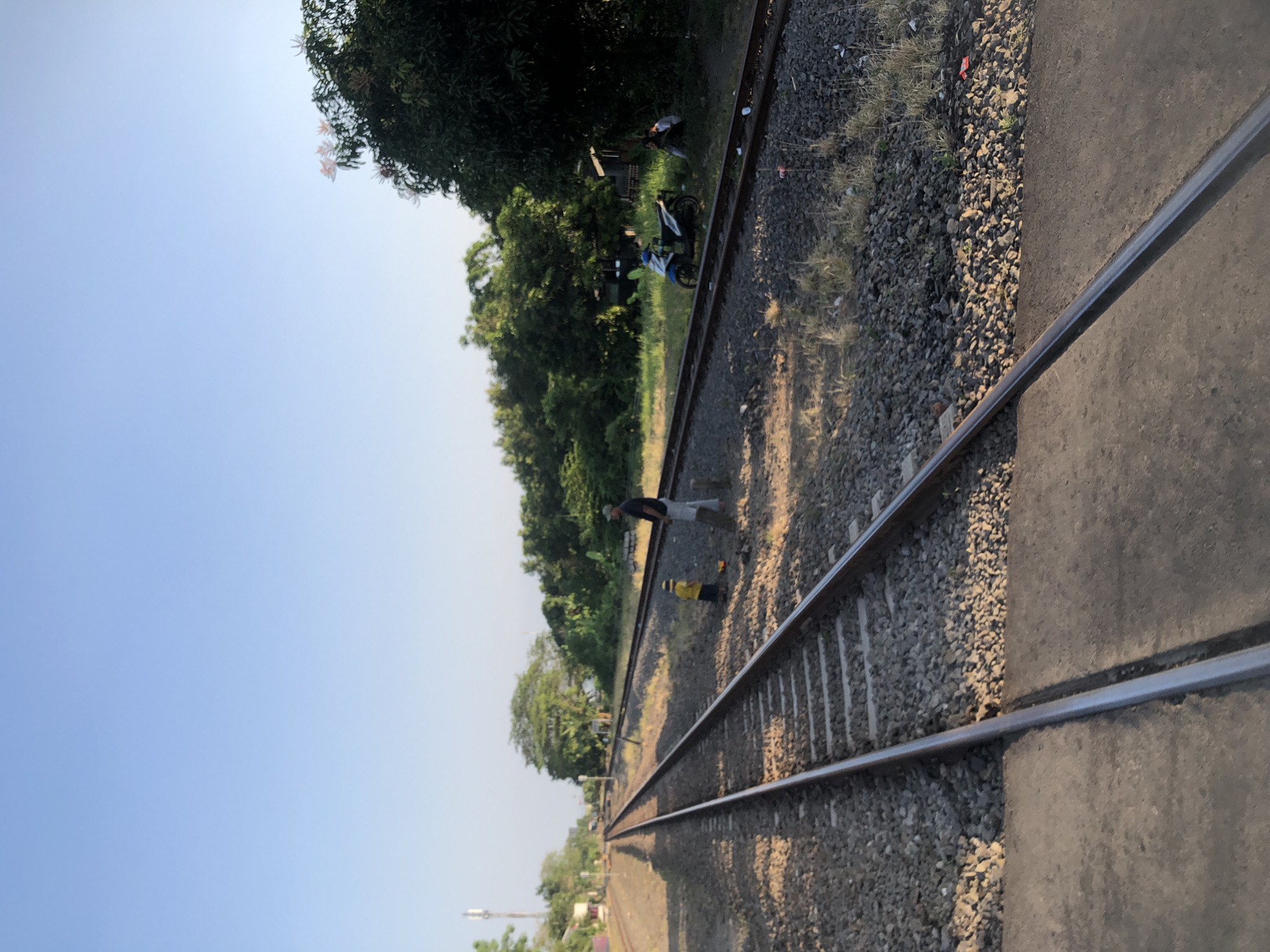 In the city of Kludan, Indonesia, a train track stretches into the distance under a clear blue sky. The gravel bed of the track is lined with ballast stones, and concrete crossings punctuate the path. A man stands near the tracks, observing a child playing with a toy. Further down the tracks, two motorcycles are parked. In the background, lush green trees and vegetation create a natural backdrop, with a few structures visible amidst the foliage. The scene depicts a serene rural setting with a touch of everyday life.