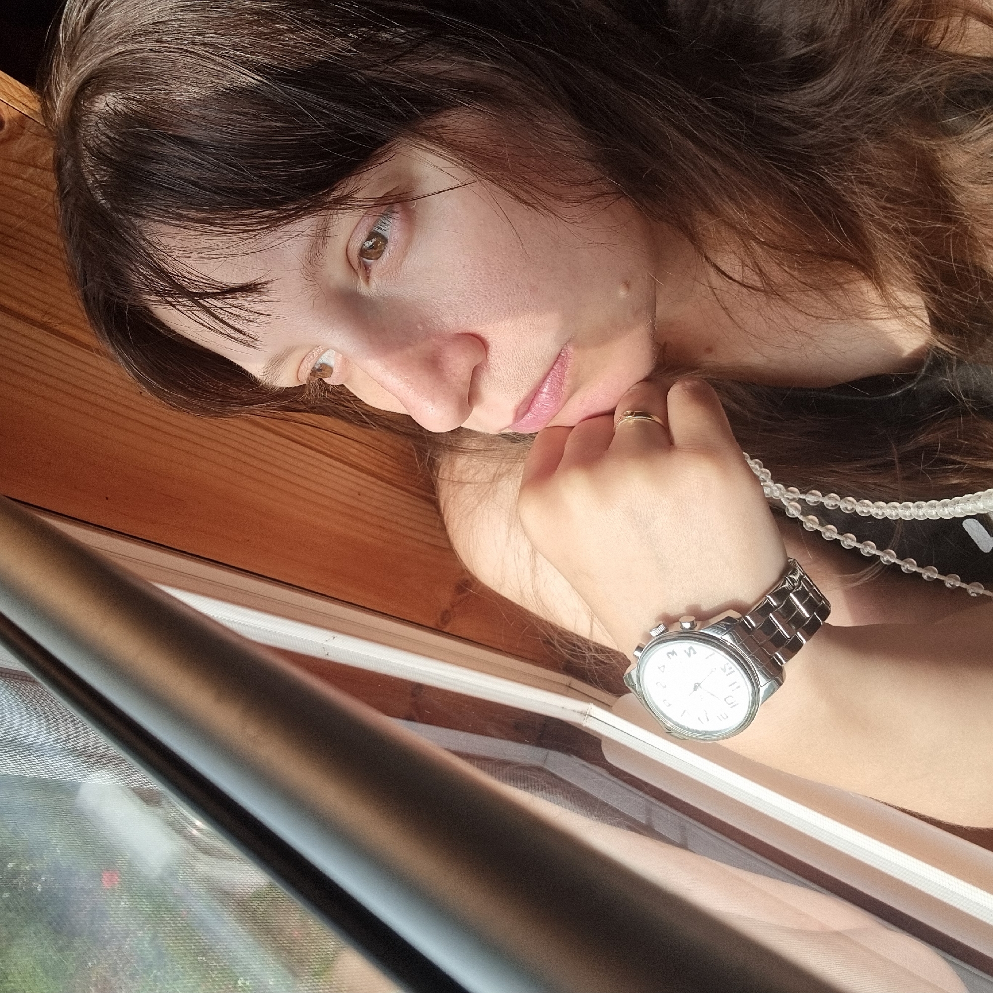 A young woman with long, dark brown hair is looking out a window. She is leaning on her hand, which is propped on her chin. She is wearing a silver wristwatch on her left wrist and a thin gold ring on her right ring finger. She appears to be in a room with wooden walls and a window that looks out onto a green, outdoor area.