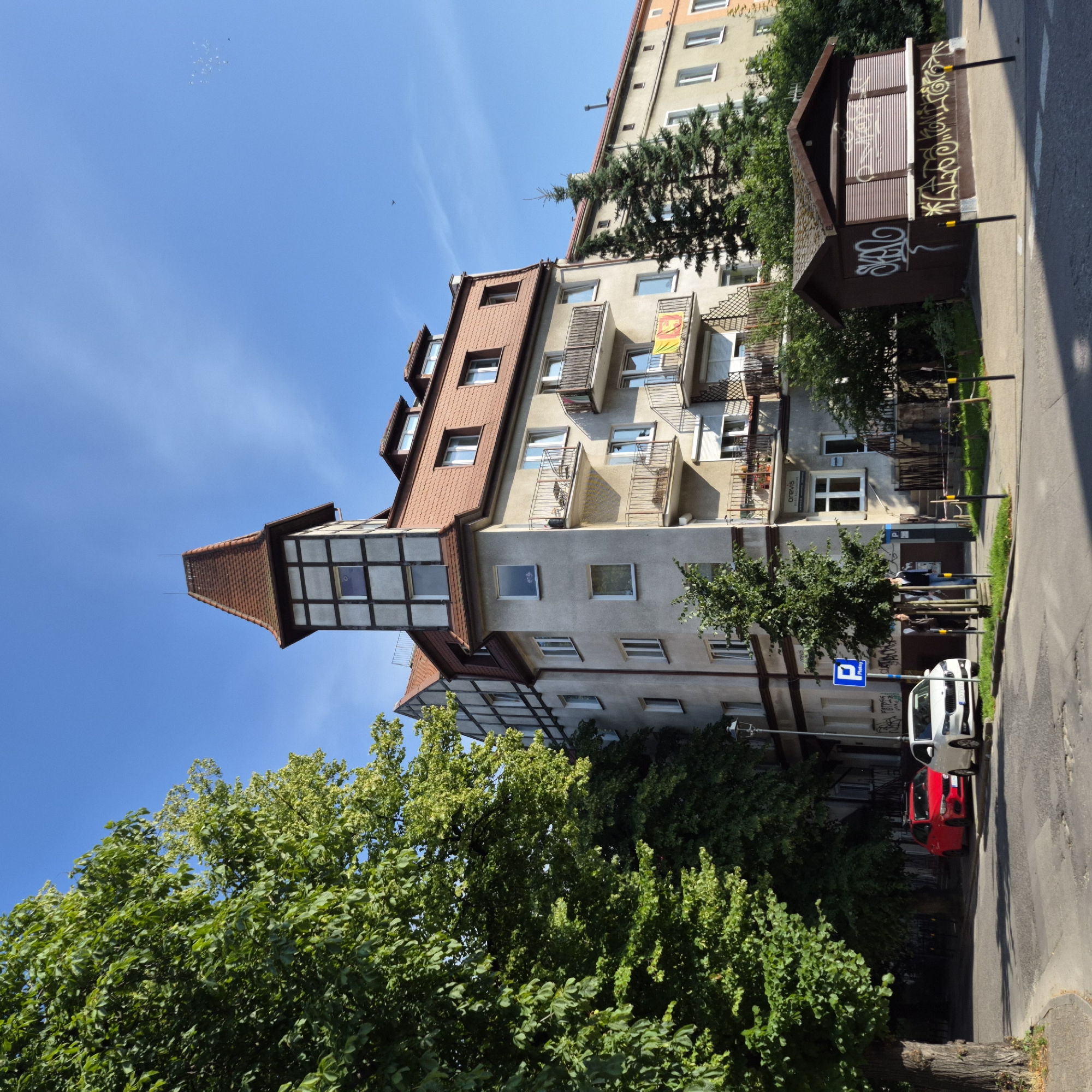 A sunny day in Gdańsk, Poland, illuminates a street scene with a multi-story building and parked cars. A large deciduous tree with dense green foliage dominates the left side of the frame, casting shadows on the asphalt road. In front of a light-colored, multi-story building with a red-tiled roof and distinctive half-timbered upper sections, a red car and a white car are parked. The building features several balconies with dark metal railings. To the right, a small, dark brown kiosk with slatted shutters is covered in graffiti. Beyond the main building, another building is visible with a similar architectural style. The sky is a clear, bright blue. Two people, a man and a woman, are walking on the sidewalk in front of the building, near a blue parking sign.