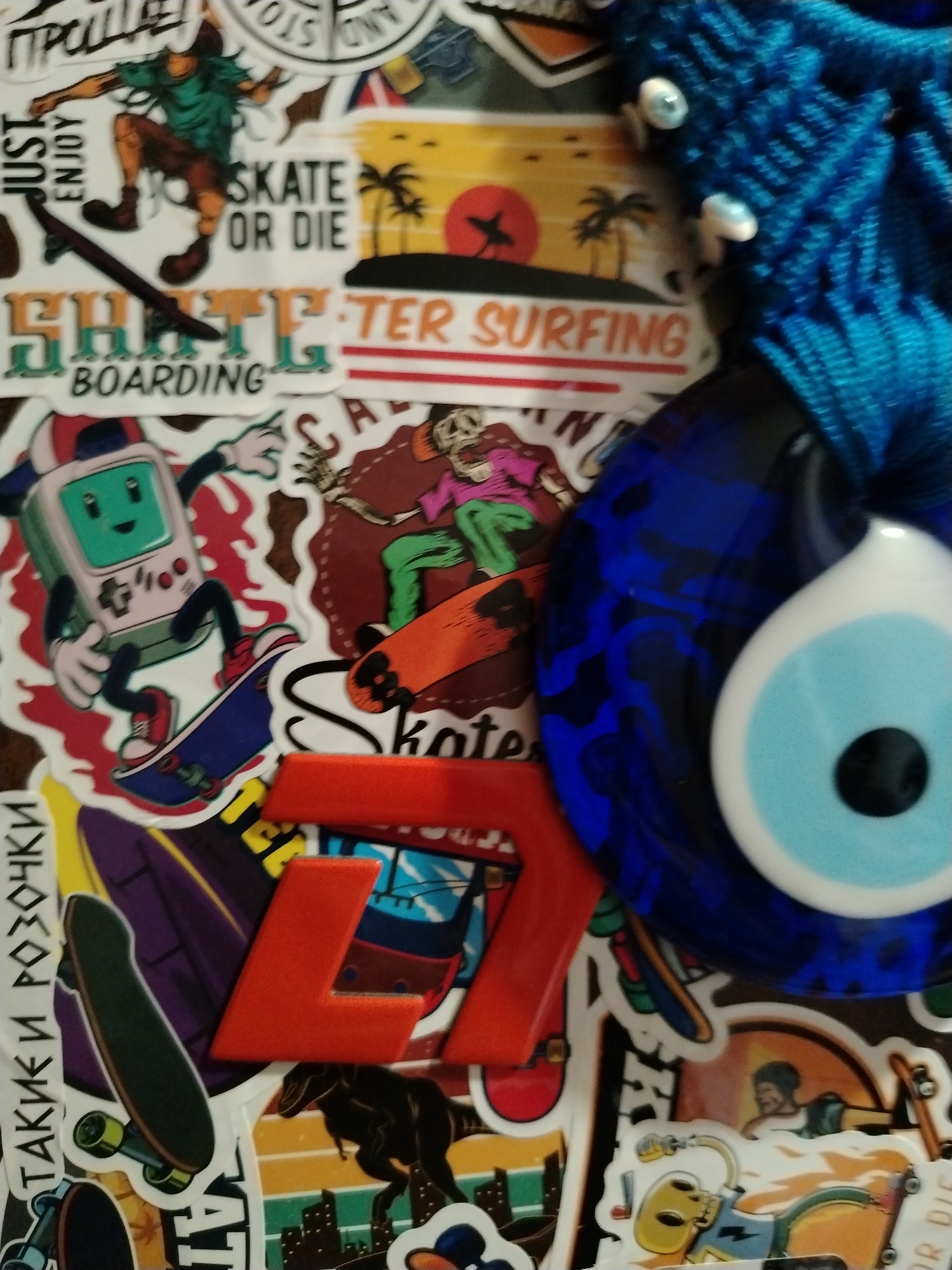 The image shows a close-up of a collection of stickers and a blue, eye-shaped charm. The stickers feature various designs, including skateboarding themes, a skeleton, a game console character, and a beach sunset. Some of the text visible on the stickers includes "SKATE OR DIE," "SURFING," and "BOARDING." The charm appears to be a Nazar amulet, commonly used for protection. The background is a dark, textured surface. No specific location in a city is identifiable.