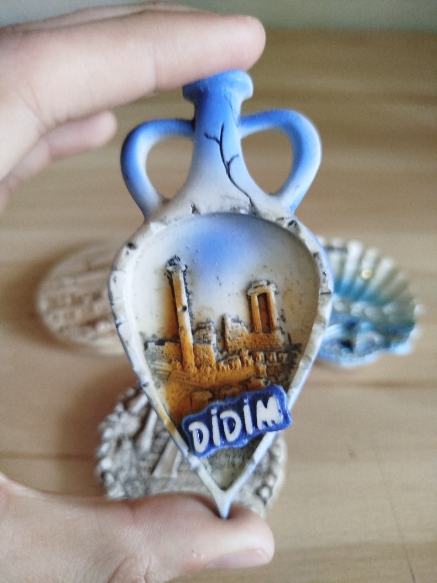 A hand holds a souvenir of Didim, Turkey. The souvenir is shaped like an amphora and depicts ruins. In the background, out of focus, are other souvenirs that are most likely on a shelf.