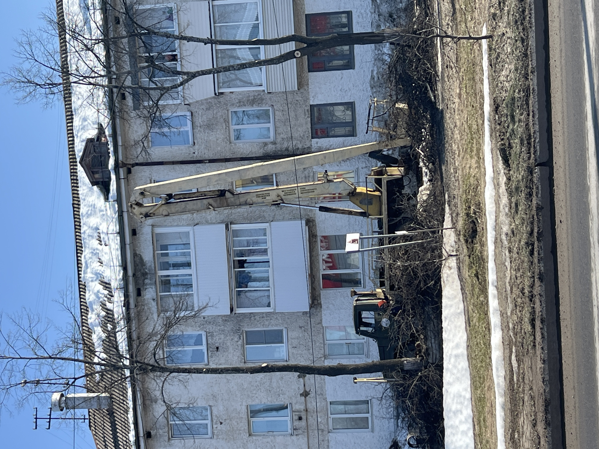 The image depicts an exterior scene in Chaikovsky, Russia, during what appears to be late winter or early spring. A light-colored, multi-story residential building with a textured facade occupies the background. Its tiled roof is partially covered with snow, featuring a chimney with an antenna structure and a small wooden dormer.
In front of the building, a large yellow mobile crane with an extended boom stands upright, positioned near the structure. To its left, partially obscured by debris, is a dark green utility truck with an open bed. The ground directly in front of the building, around the equipment, is covered with extensive piles of cut tree branches and brush. Several bare deciduous trees are visible, one prominently in the foreground, showing evidence of recent trimming.
Patches of snow are present on the ground, interspersed with areas of dry, light-colored grass and exposed earth. A paved road or street with a curb runs across the immediate foreground. The sky above is clear and blue. One individual is faintly visible peering from a second-story window of the building, to the right of the crane's boom. No other explicit human activity or interaction is clearly discernible.