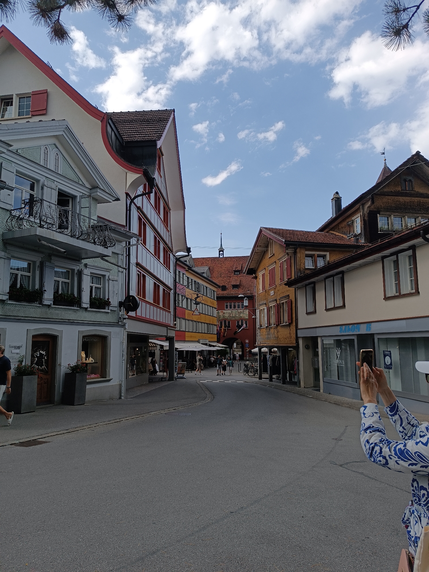 A street scene in Appenzell, Switzerland, is depicted on a sunny day. Buildings line both sides of the street, with a mix of architectural styles. On the left, a light-colored building with red shutters features a balcony with decorative metalwork. To its right, a building with a red and white facade has multiple windows. Further down the street, buildings display yellow and red facades with traditional shutters. The street is paved and appears to be a pedestrian area, with a few people walking and some outdoor seating for cafes. In the foreground, on the right, a person is holding up a smartphone, seemingly taking a picture of the street. The sky is blue with scattered white clouds. A branch of a pine tree is visible in the upper left corner of the image.