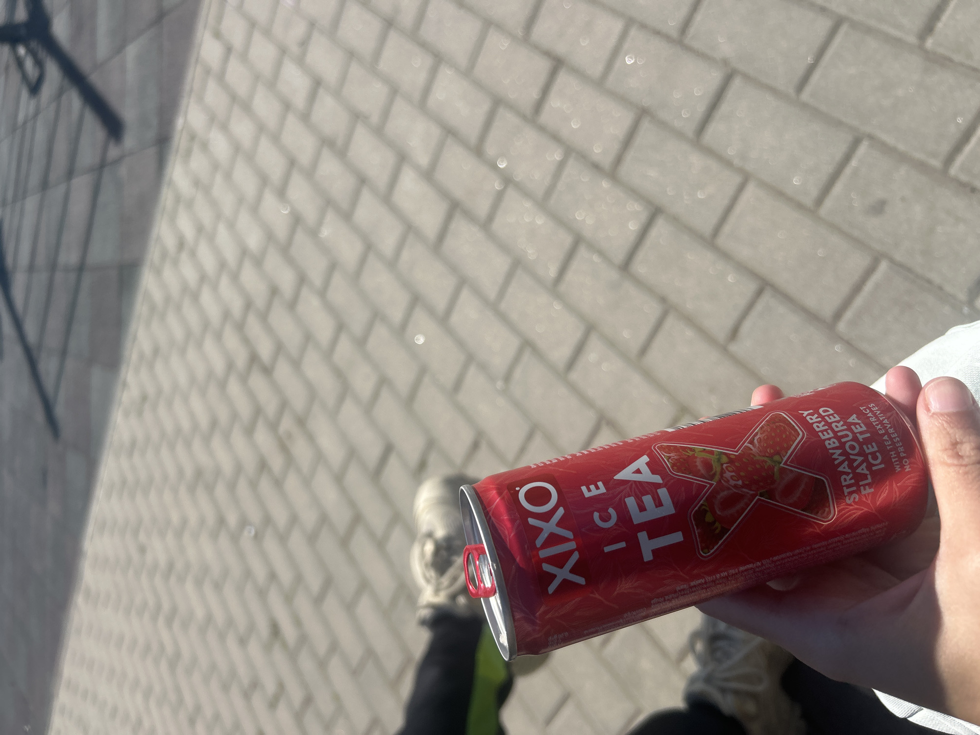 The image shows a hand holding a can of "Xixo Ice Tea" with strawberry flavor. The background reveals a paved walkway, suggesting an urban setting. The person holding the can is likely standing on the walkway, with their legs and shoes partially visible in the lower part of the image. The lighting suggests a sunny day. It's impossible to confirm the exact location in Burgas, Bulgaria, without more specific contextual clues.