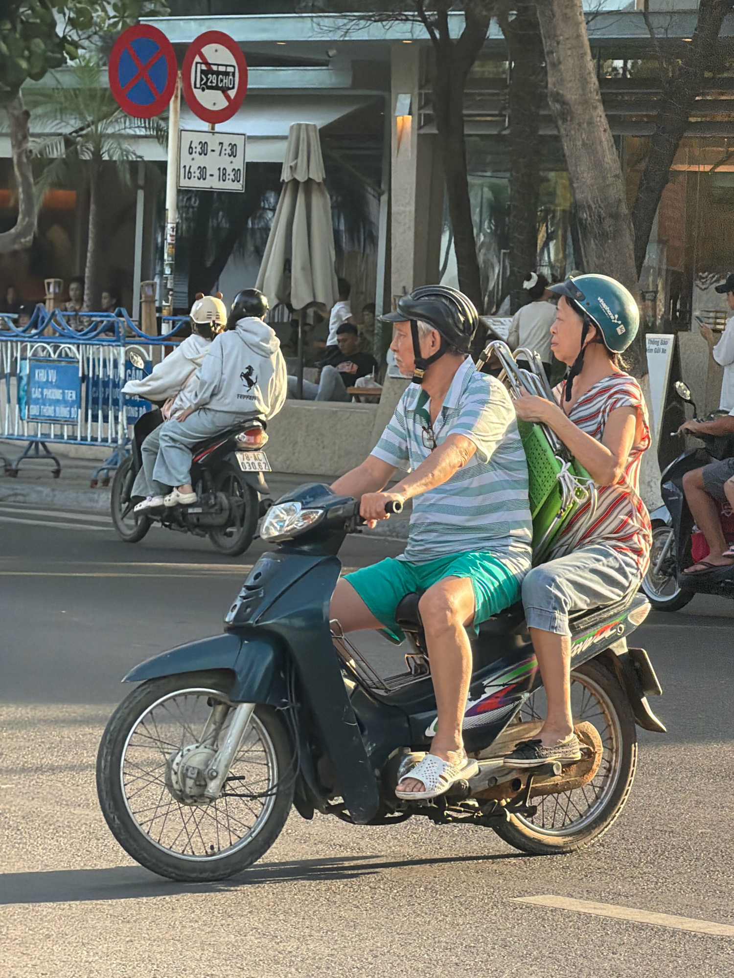 The image captures a street scene in Nha Trang, Vietnam, specifically on Tran Phu Street, near a busy intersection. In the foreground, a man and a woman ride a dark-colored scooter. The man, wearing a striped t-shirt, teal shorts, and a black helmet, is driving. The woman, in a striped t-shirt and denim pants, wears a green helmet and is holding a large green bag. Behind them, another scooter carries two people wearing casual clothing and helmets.
The background reveals the urban environment of Nha Trang. Several traffic signs are visible, including a circular red sign with a blue cross, indicating no parking, and another circular red sign with a bus icon and the text "29 CHO," suggesting restrictions for vehicles carrying 29 passengers. Below these, a rectangular sign displays time restrictions: "6:30 - 7:30" and "16:30 - 18:30". Buildings with large windows, awnings, and trees line the street. In the distance, more vehicles and pedestrians can be seen, indicating a bustling city atmosphere. The lighting suggests it is late afternoon, with the sun casting long shadows. The road surface is asphalt.