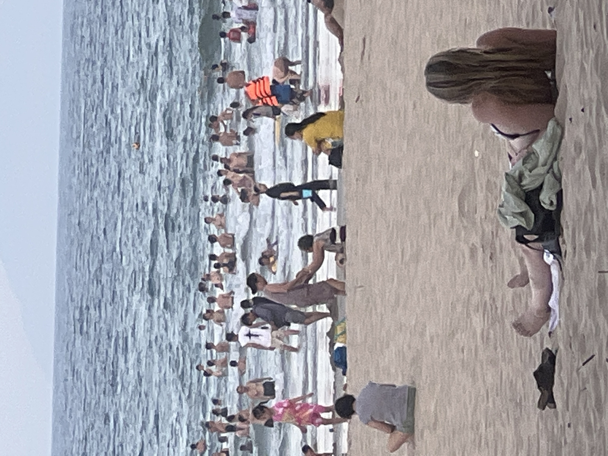 The media file depicts a crowded beach scene in Đà Nẵng, Vietnam. The primary subjects are numerous people enjoying a day by the sea, with many visible in the water and on the sandy shore.
In the water, people are engaged in various activities, including swimming and wading. Some are seen closer to the shore, while others are further out. One individual in the water is wearing a bright orange life vest.
On the sandy beach, more people are gathered. A woman with long blonde hair is prominently featured in the foreground, facing away from the camera, sitting on the sand. She is wearing a dark swimsuit and has a light-colored garment draped around her. Nearby, a person in a grey t-shirt and shorts is crouched on the sand. Further along the beach, a group of people are interacting, some appearing to be in motion. A child in a pink patterned outfit is visible, and another person in a white t-shirt with a cross emblem is also present. The sand appears to be a light beige color.
The overall atmosphere suggests a lively and busy day at the beach, likely during daylight hours, given the presence of sunlight. The water is a medium blue, with waves visible. The sky is a light grey, indicating potentially overcast or hazy conditions. The location is clearly identified as Đà Nẵng, Vietnam, a popular coastal city. No visible text is present in the media file.