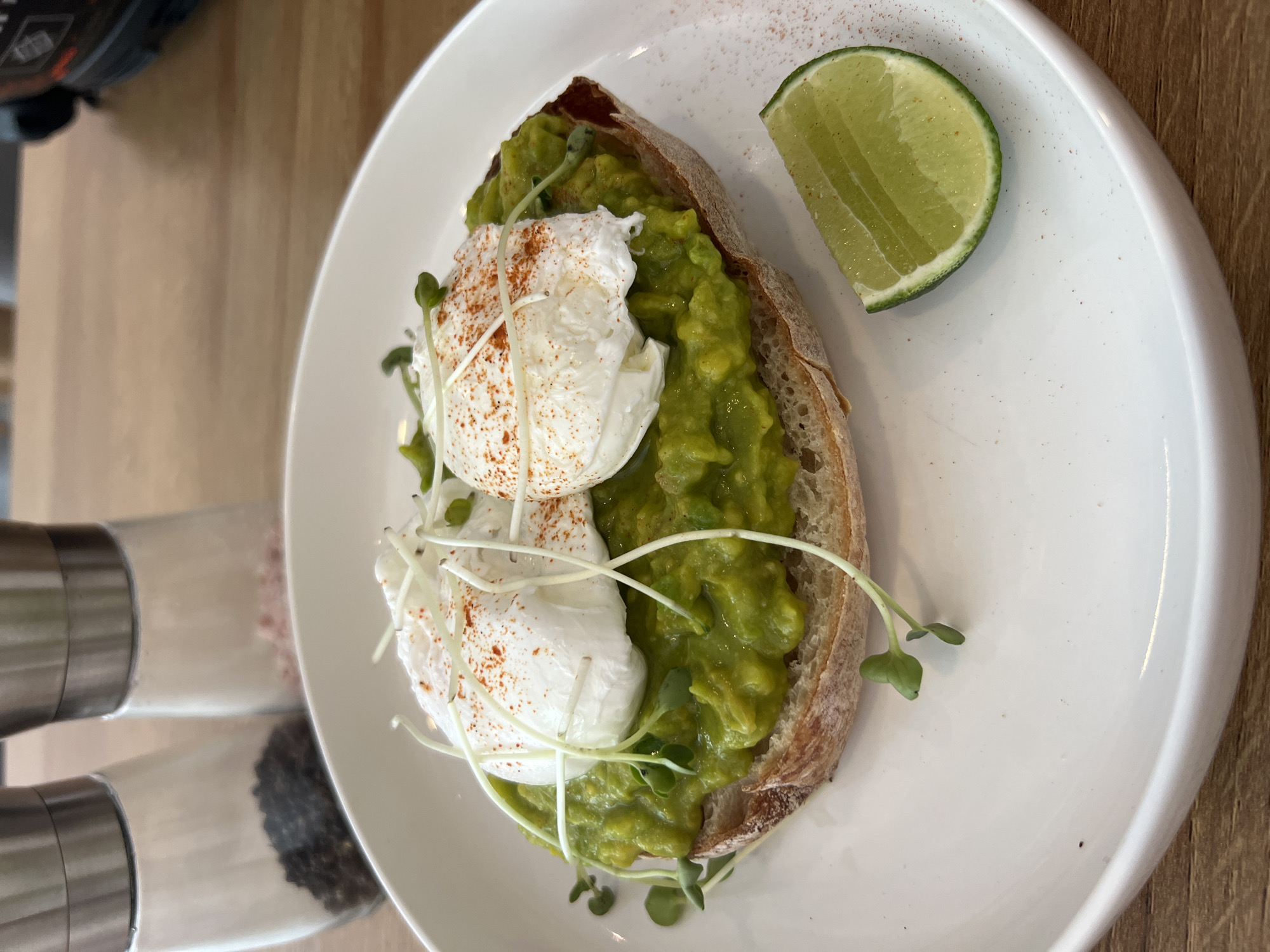 A top-down shot depicts avocado toast on a white plate, with a lime wedge on the side. The toast is made of bread that is topped with mashed avocado. Two poached eggs, sprinkled with a reddish powder (possibly paprika), sit atop the avocado. Some sprouts are added as garnish. To the side of the plate are two salt and pepper shakers. The scene is located indoors, on a wooden table or surface. The location in the city is Unknown, Unknown.