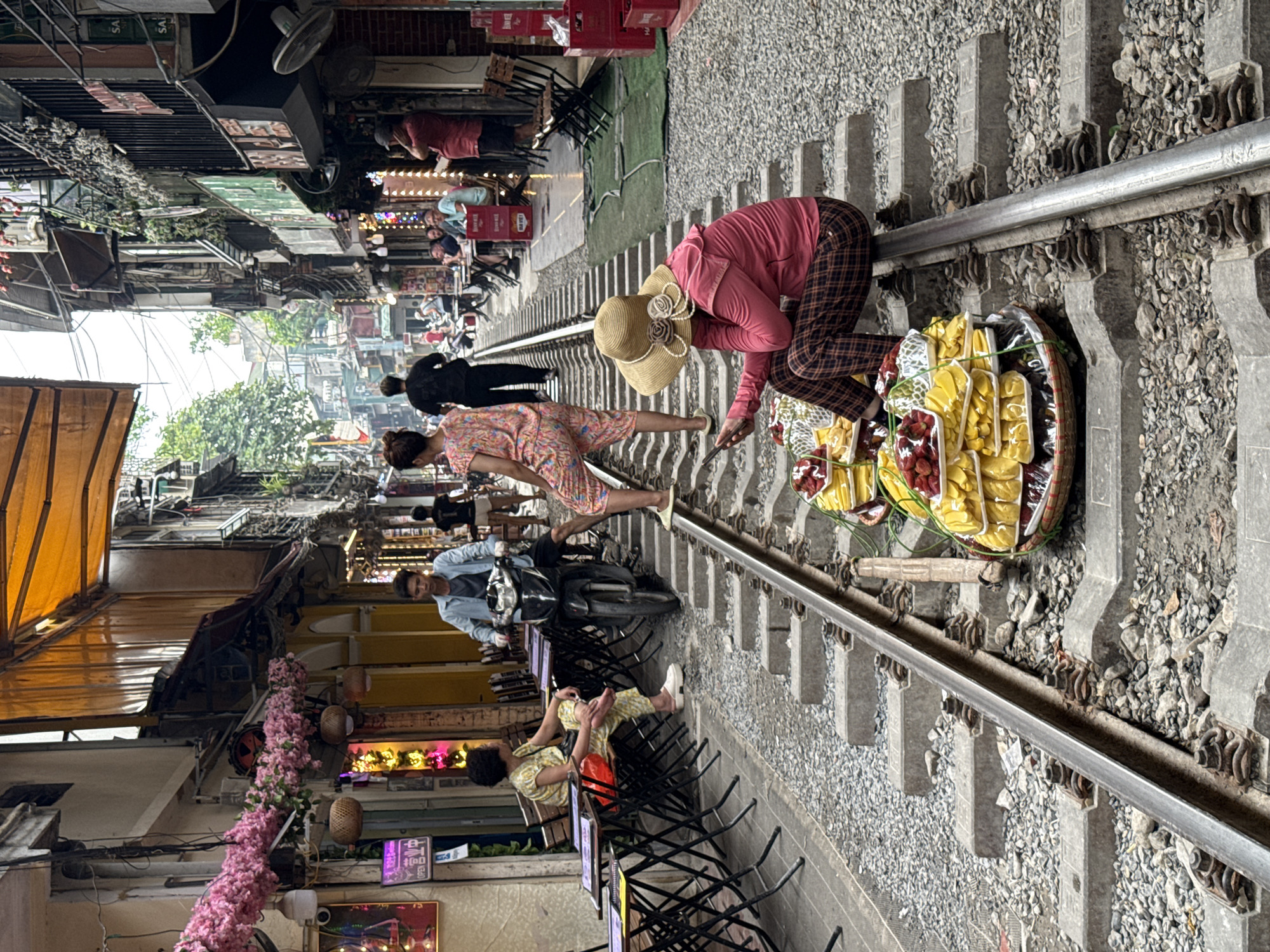 This detailed image captures a bustling street scene along the active railway tracks of Hà Nội, Vietnam, commonly known as "Train Street." The single set of tracks, composed of metal rails on concrete sleepers and ballast, bisects a narrow urban corridor flanked by multi-story buildings that serve as residences and commercial establishments, primarily cafes and shops.
In the immediate foreground, positioned on the right rail, a vendor sits wearing a pink long-sleeved shirt, brown and black plaid pants, and a wide-brimmed straw hat adorned with flowers and pearls. This individual is looking down at a smartphone held in their left hand. Adjacent to them, on the tracks, are two large, round, woven baskets overflowing with pre-packaged fruits, including bright red strawberries and stacks of sliced yellow fruit, likely mango or jackfruit.
Moving into the mid-ground along the tracks, a person in a light blue long-sleeved shirt and dark pants is operating a black scooter, riding on the tracks and facing towards the viewer. Directly behind the scooter rider, a person in a pink, white, and orange floral jumpsuit stands with their left foot on a concrete sleeper and their right foot raised onto the rail. Further back on the tracks, a person in a dark t-shirt and dark shorts walks away from the viewer.
To the left of the tracks, several collapsible metal tables and chairs form an outdoor cafe setup. An older woman with curly dark hair, dressed in a yellow patterned outfit, sits at one of these tables, facing right with her left leg crossed. Additional patrons are visible seated at other tables along both sides of the railway further down the street. On the right side, a man in a red t-shirt and dark shorts stands near a shop entrance, next to stacked red plastic crates.
The buildings lining the street feature various architectural details, including balconies, windows, and ground-level storefronts. An orange fabric awning extends from a building on the left, adorned with clusters of artificial pink flowers and colorful neon lights. The sky overhead is bright and overcast. The overall scene illustrates a unique blend of daily life and commerce integrated with the active railway environment.