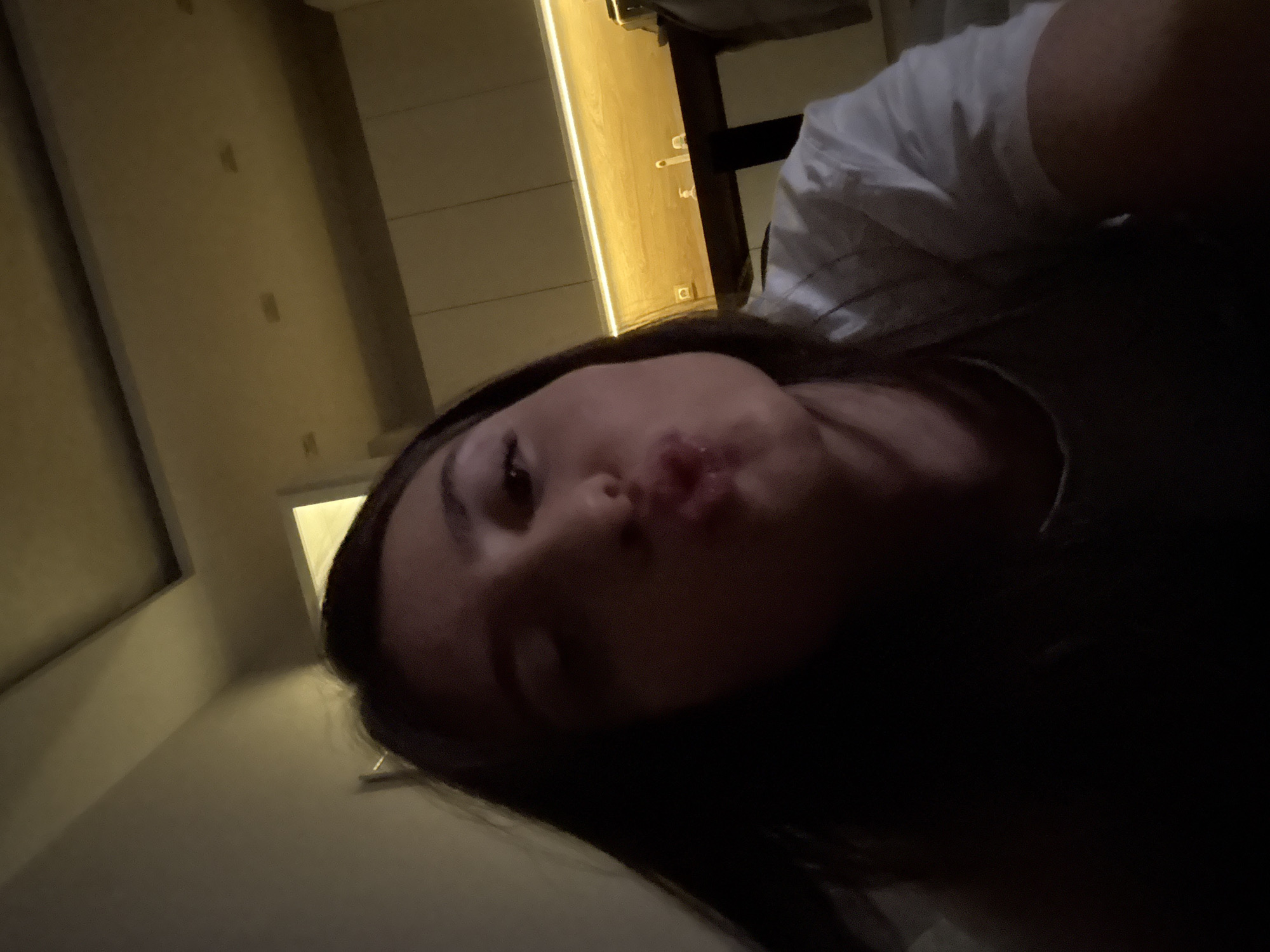 A dimly lit indoor shot, possibly taken at night, features a young woman with dark hair as the primary subject. Her face is close to the camera, and she appears to be making a kissing face or pouting. She is wearing a light-colored shirt, possibly white.
The background is blurred but appears to be an interior space, with what may be a door or closet on the right side and a wall on the left. The location in Plovdiv, Bulgaria, is unknown.